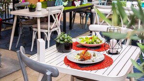 Un área de comedor al aire libre con mesas y sillas blancas, rodeada de plantas y flores en macetas.