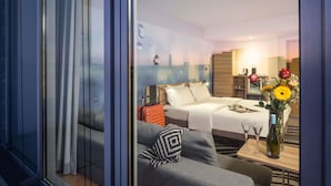 Una habitación de hotel moderna con cama, sofá, mesa de comedor con copas de vino y un florero con flores.