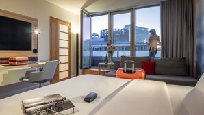 Habitación de hotel moderna con una cama grande, un escritorio con una computadora portátil, una silla, un sofá y un amplio ventanal con vista a la ciudad.