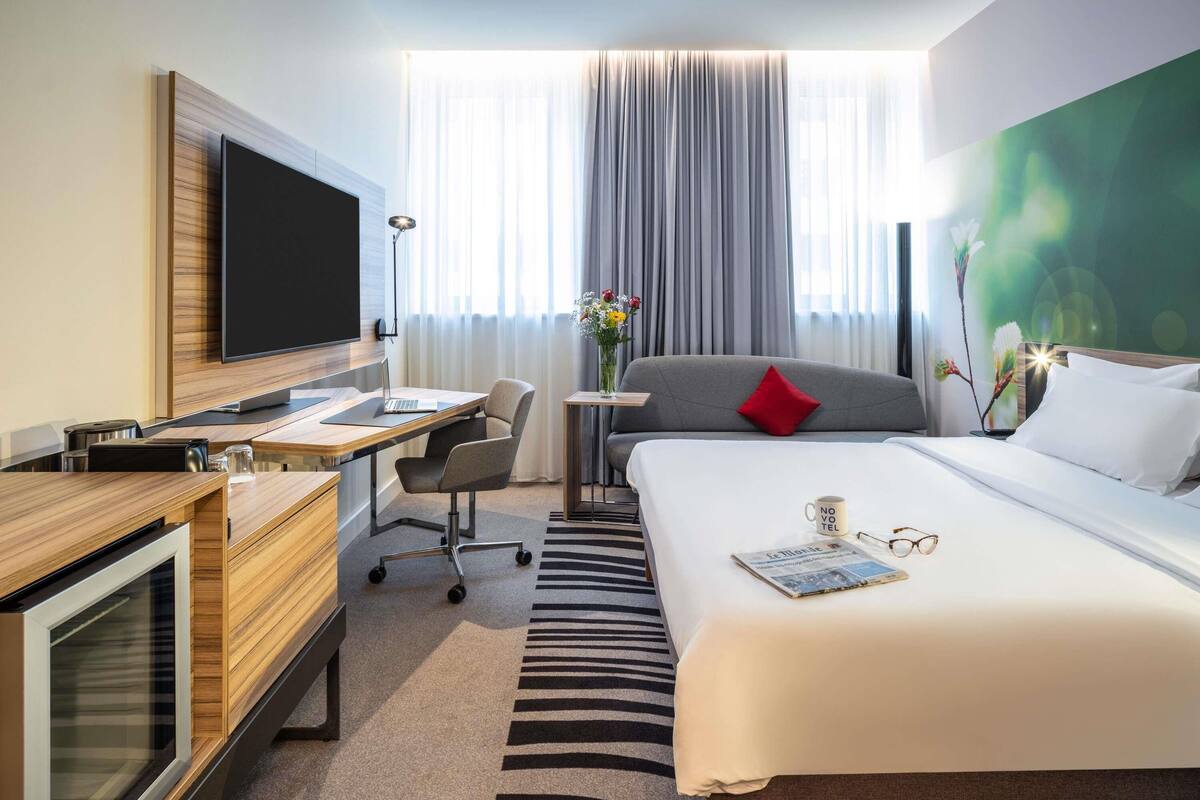 A hotel room with a bed, a desk with a computer, a TV, and a window with curtains.