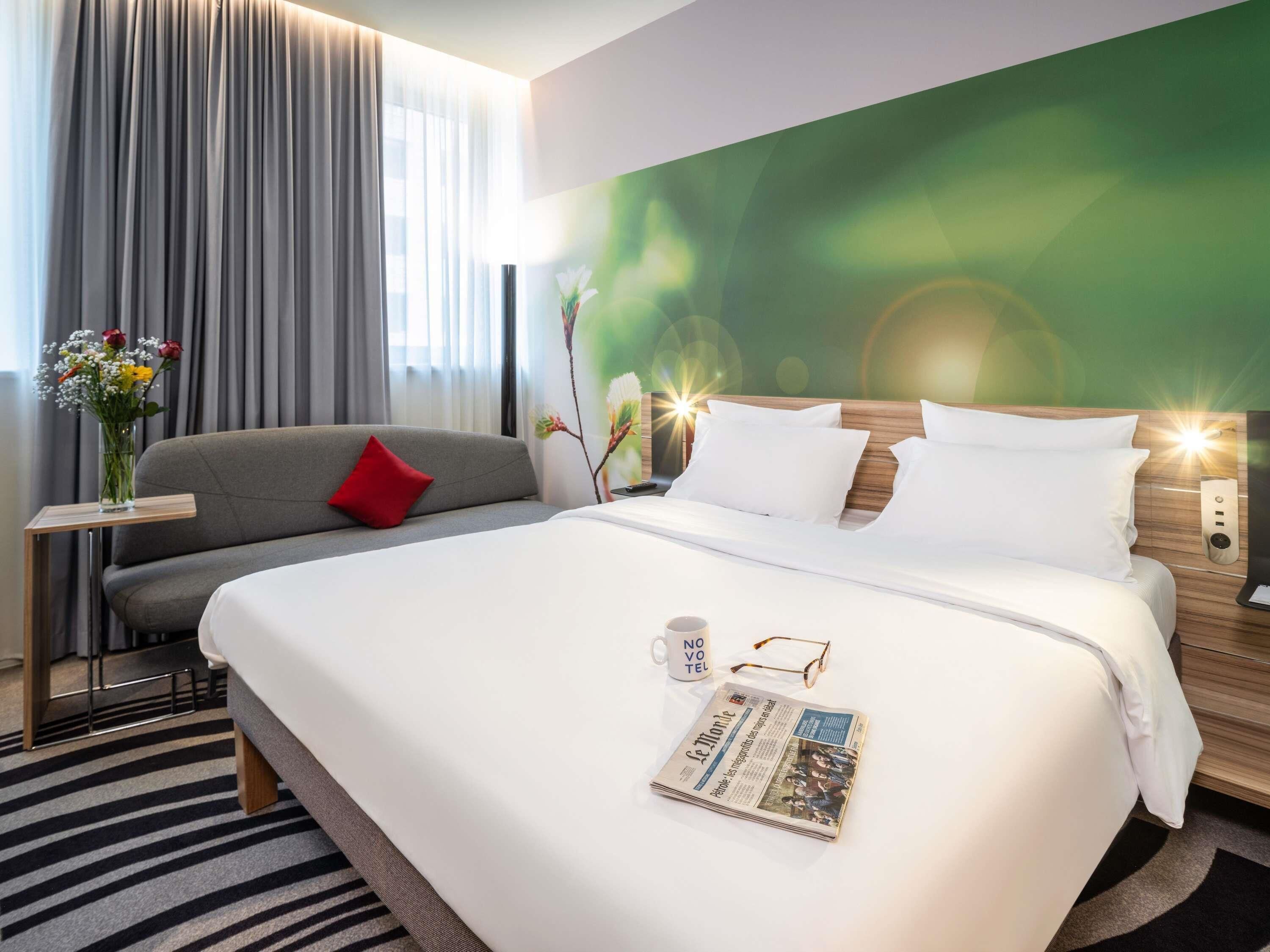 Ein Hotelzimmer mit einem großen Bett, einem grauen Sofa, einem kleinen Tisch mit Blumen und einer grünen Wand mit einer Leuchte.