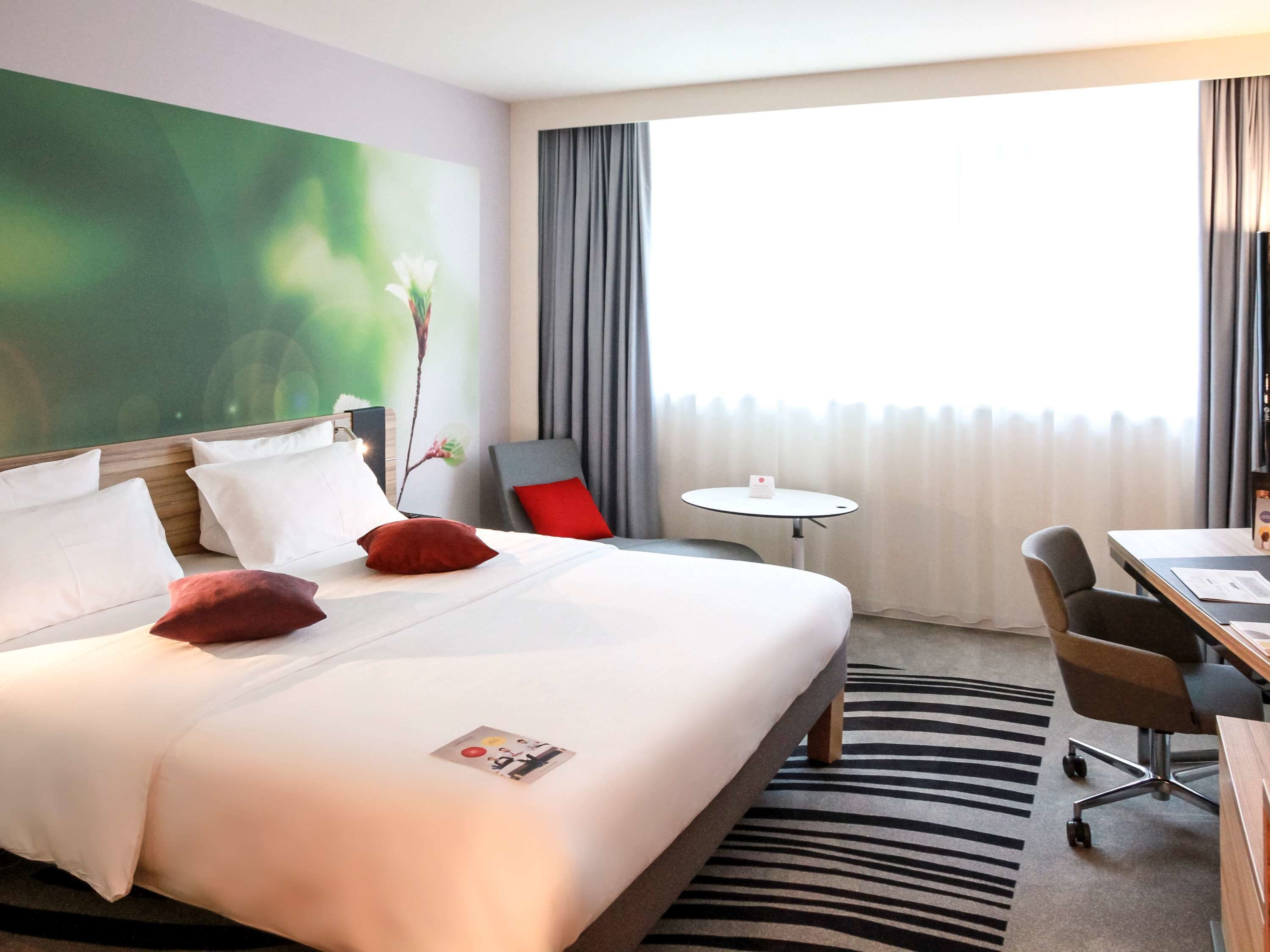 A hotel room with a bed, a desk, a chair, a small table, and a large window with curtains.