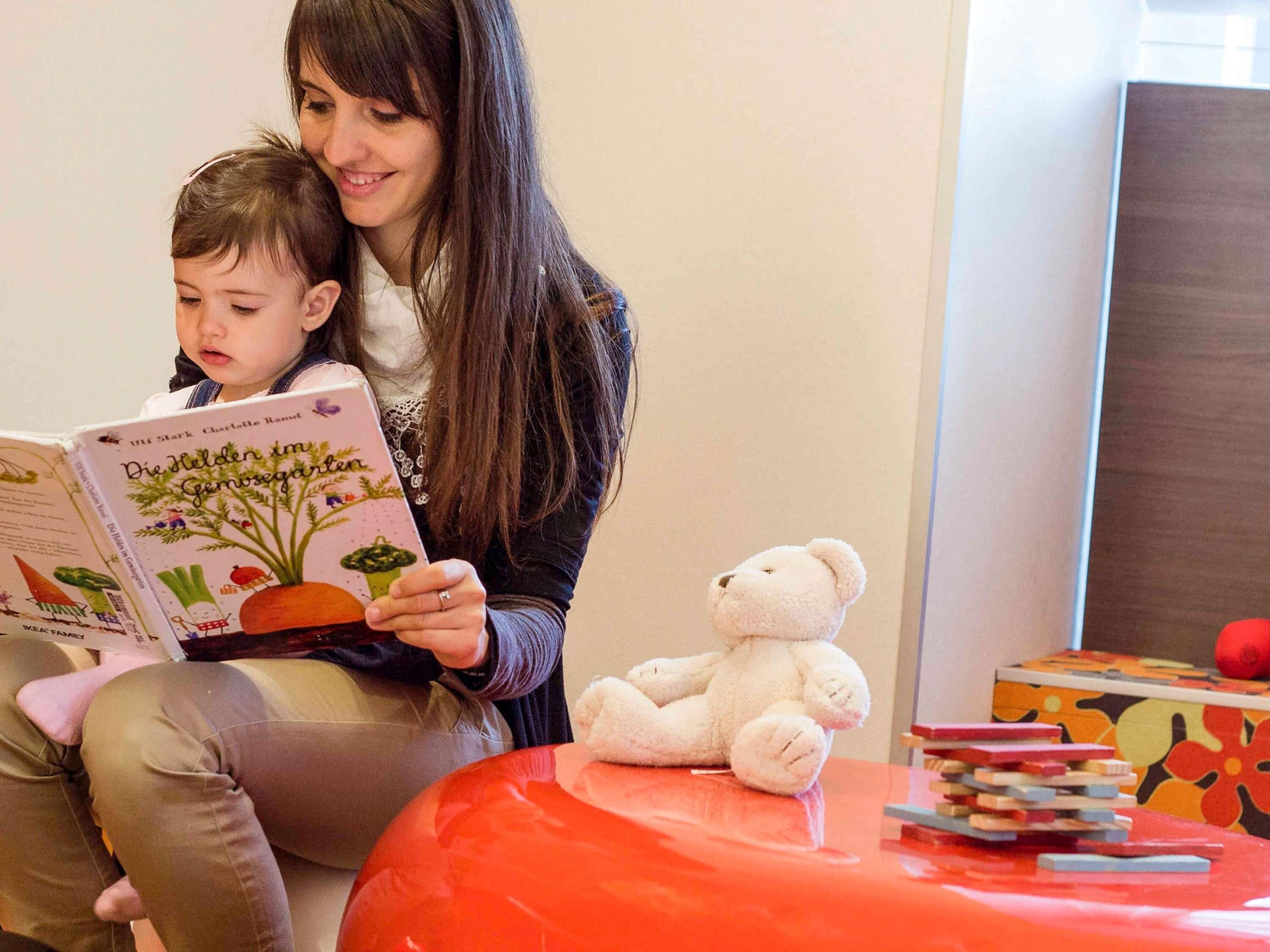A person reading a book to a child in a room with a red chair, a white teddy bear, and a stack of books.