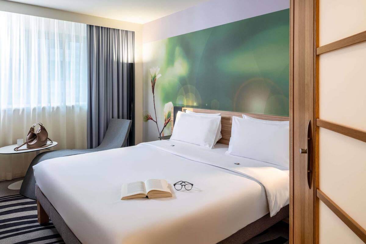 A hotel room with a large bed, a bedside table with a handbag, a chair, and a painting on the wall.