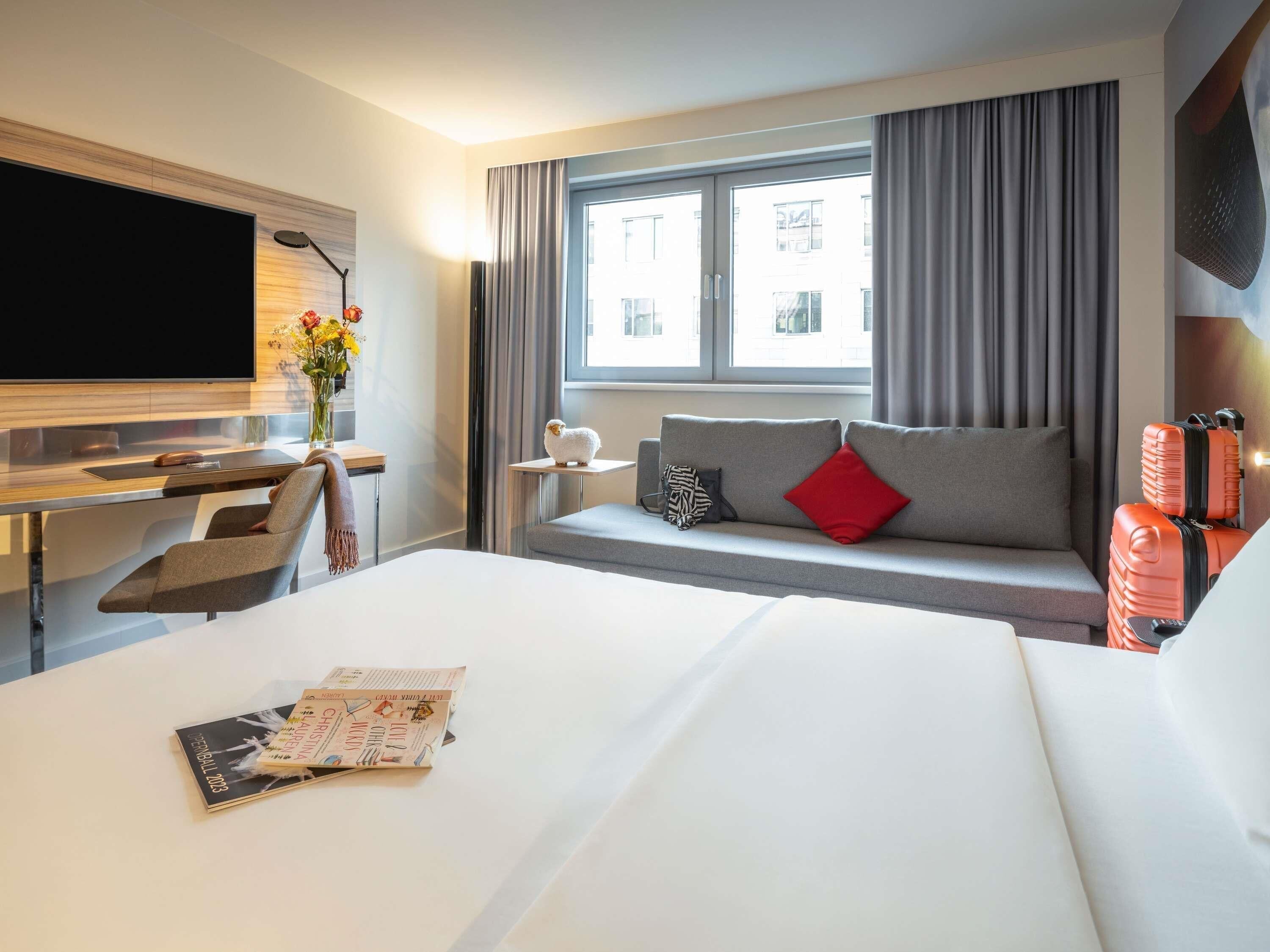 A hotel room with a bed, a desk, a TV, a window with curtains, and a sofa with pillows.