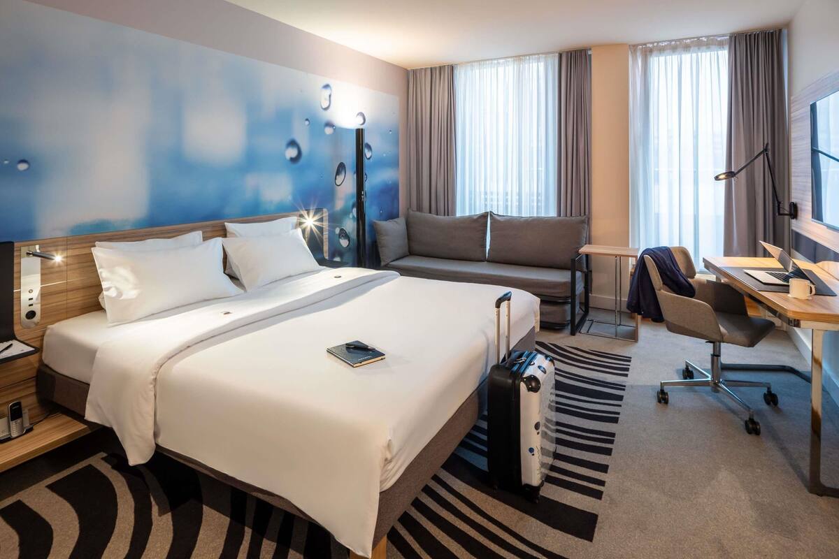 A hotel room with a bed, a desk with a laptop, a chair, a sofa, and a large window with curtains.