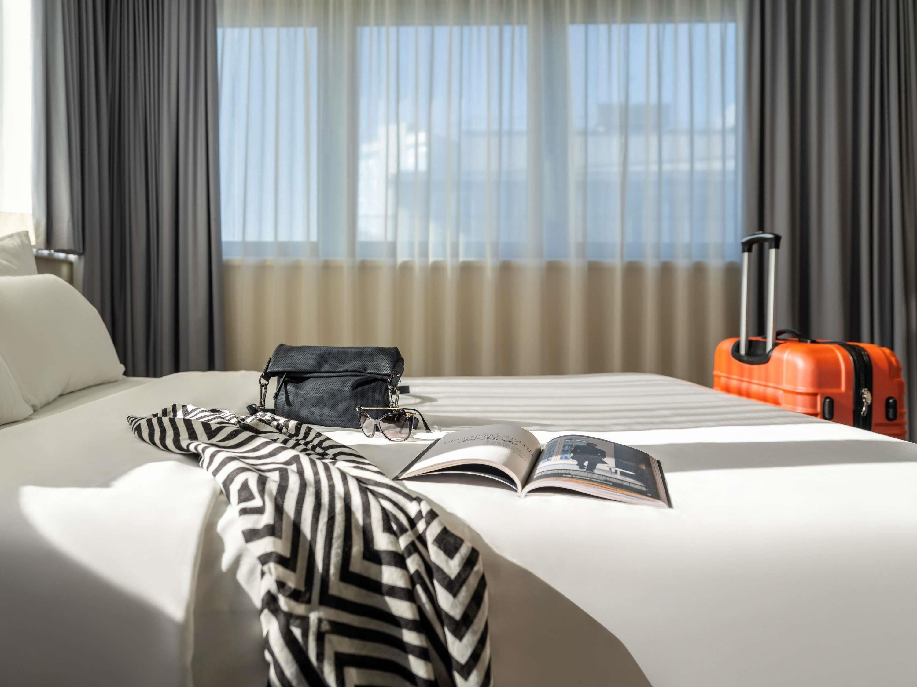 Une chambre d’hôtel avec un lit, un sac à main noir, une paire de lunettes, un magazine ouvert et une valise orange.