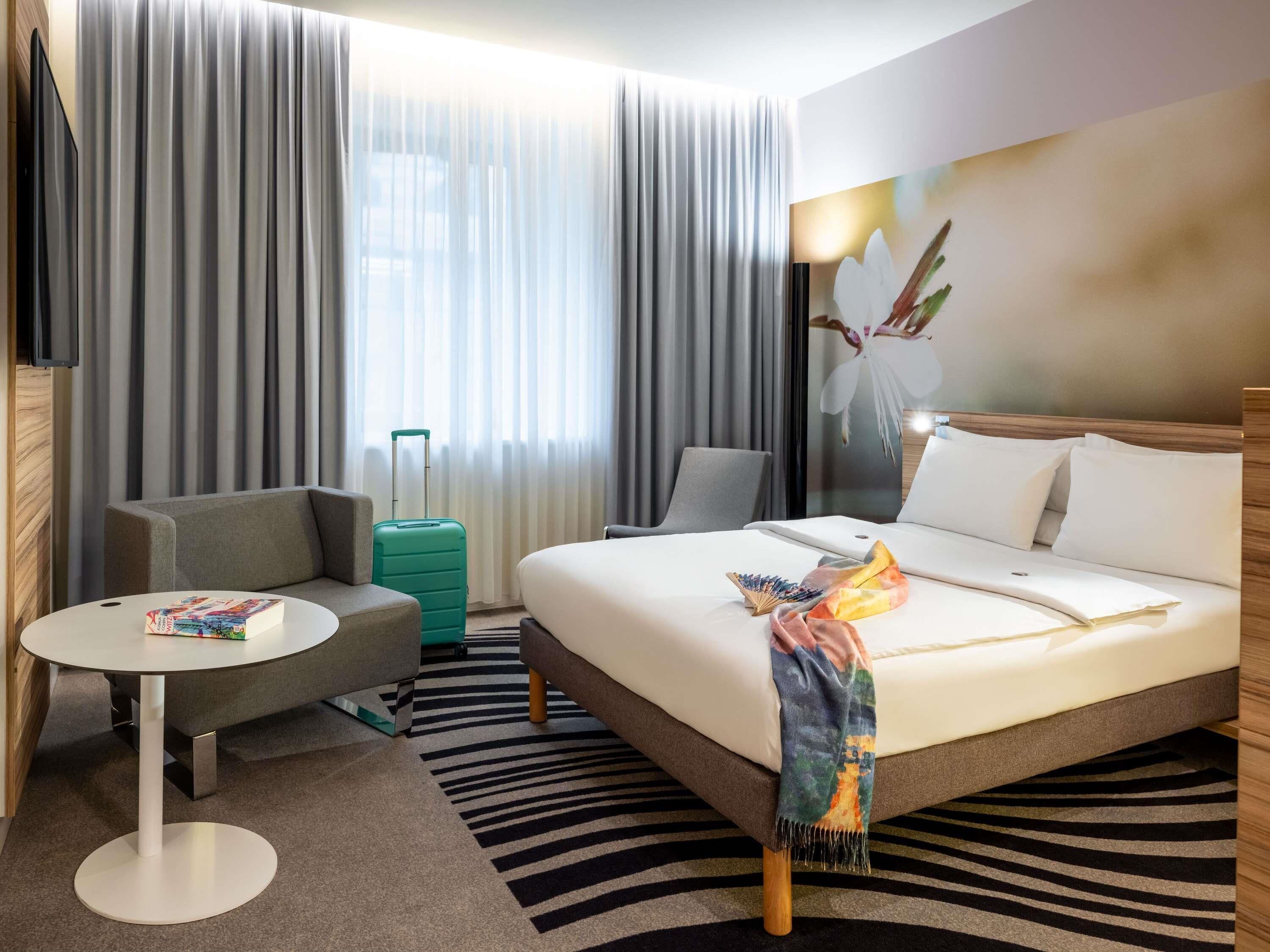 A hotel room with a bed, a desk, a chair, a suitcase, and a television.