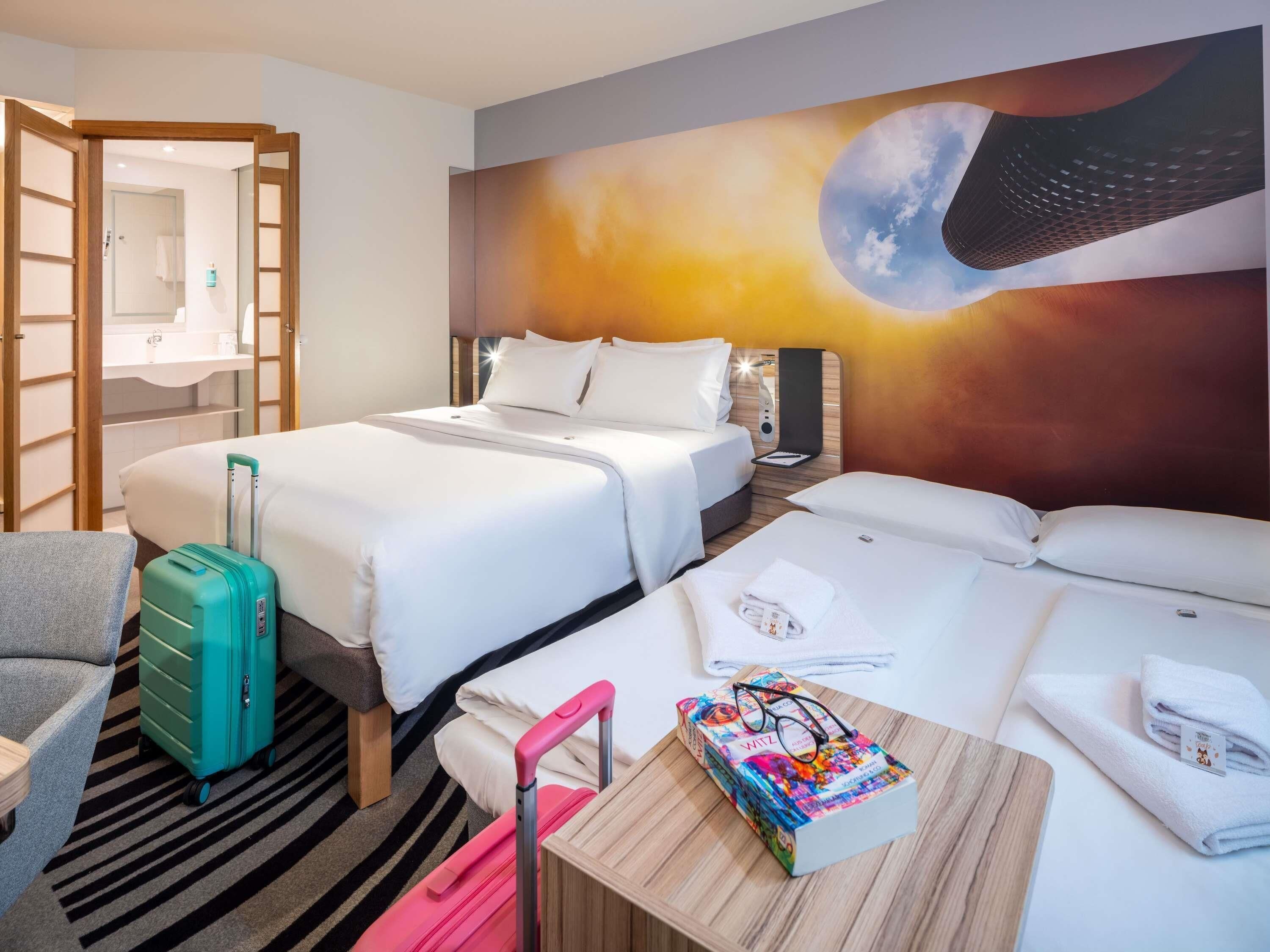 A hotel room with two beds, a desk, a chair, a suitcase, and a wall mural.