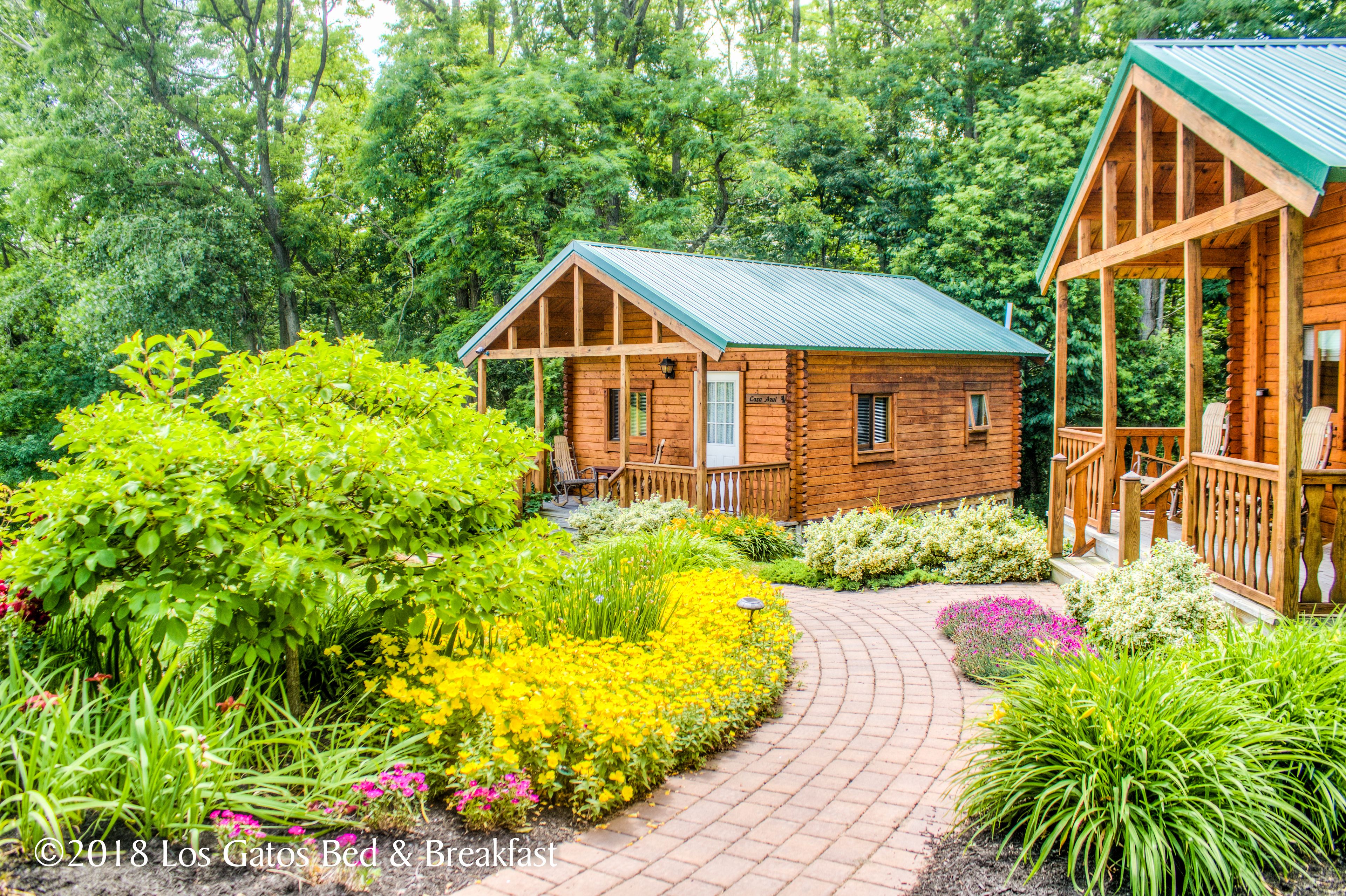 A wooden cabin with a porch and a brick pathway leading to it, surrounded by lush greenery and flowers.