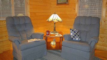 A cozy room with two grey armchairs, a wooden side table with a lamp, a small cat figurine, and a framed picture on the wall.