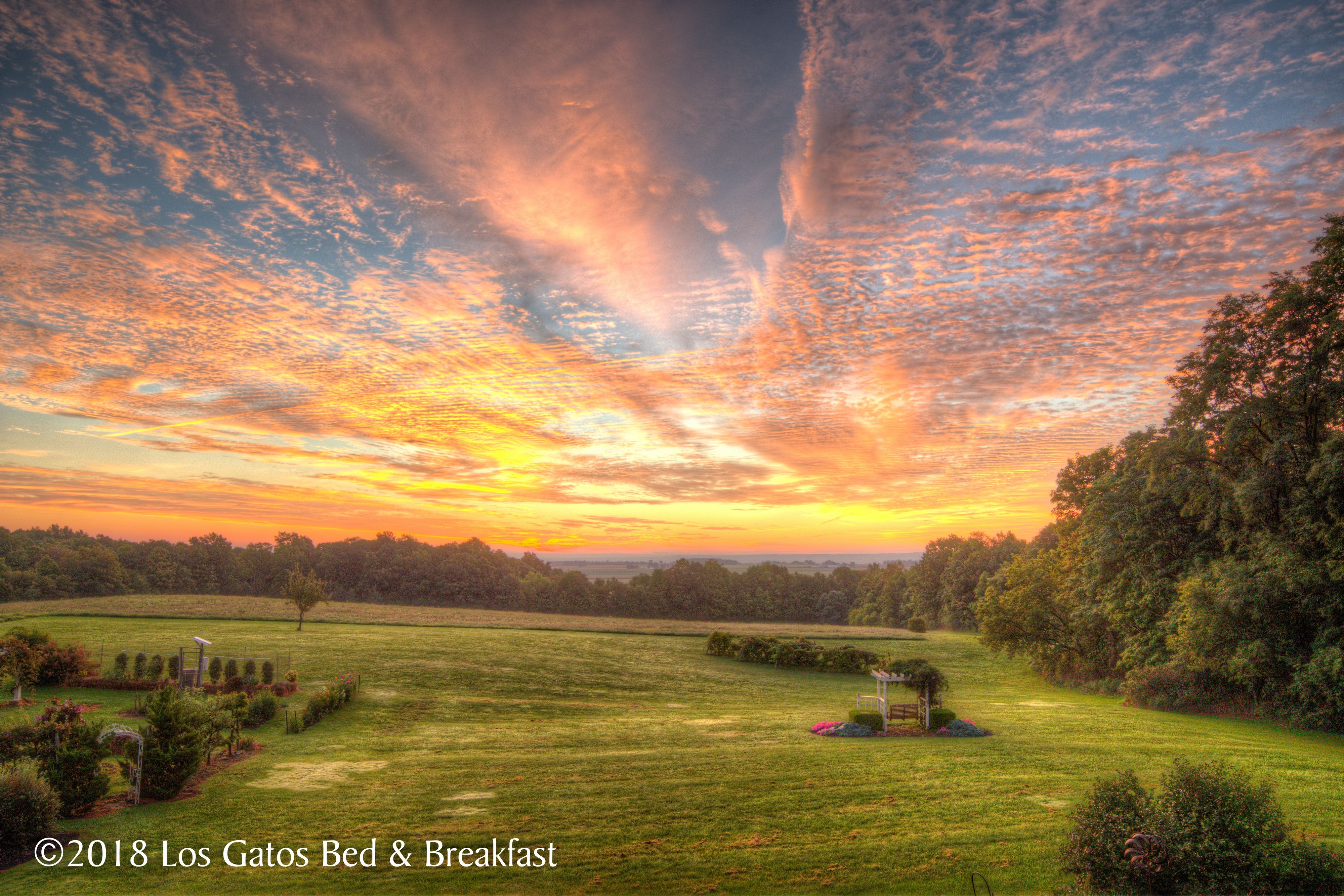 A rural landscape with a gazebo, open fields, and trees under a sunset sky.