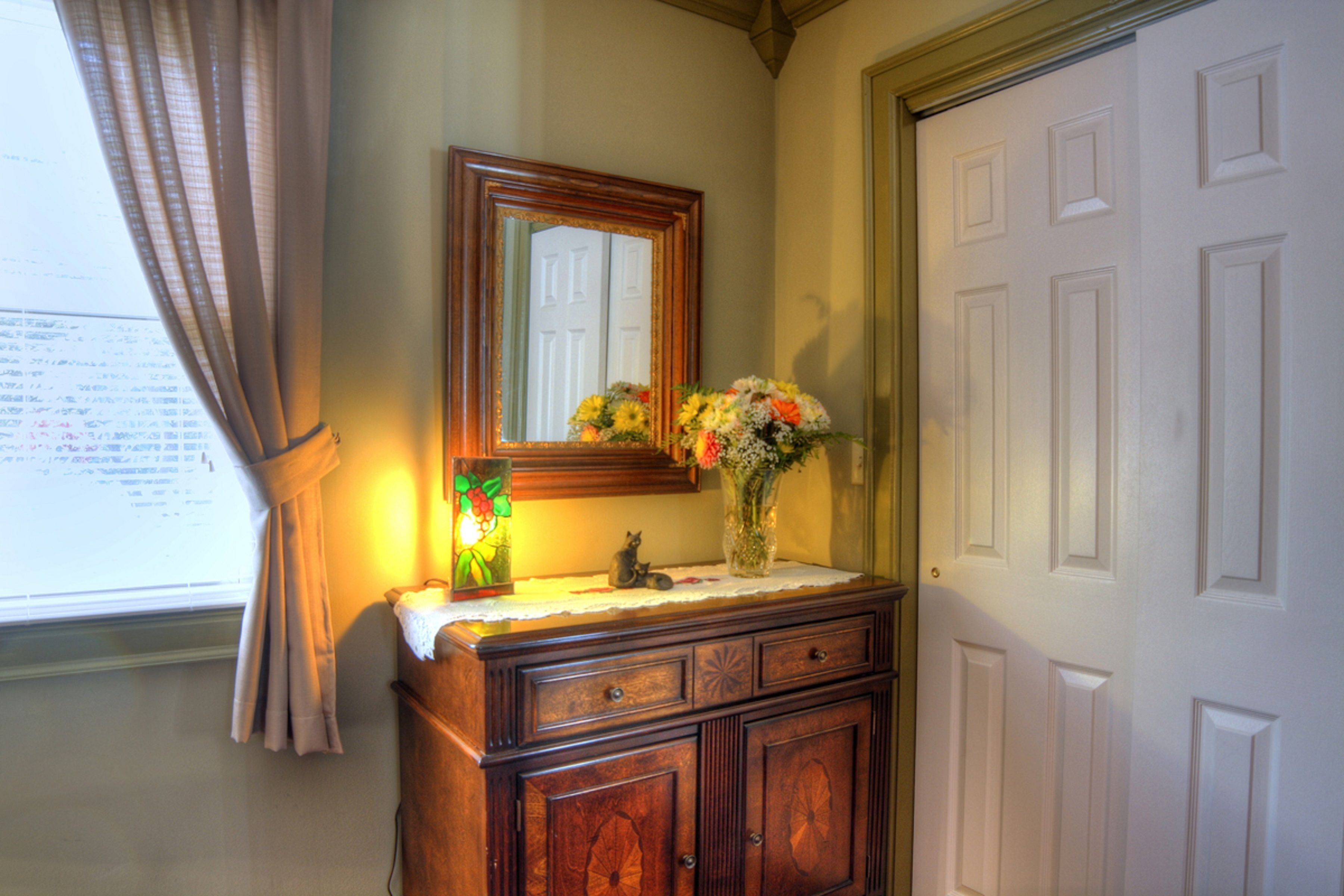 Un baño con tocador de madera, un espejo, un florero con flores y una ventana con cortinas.