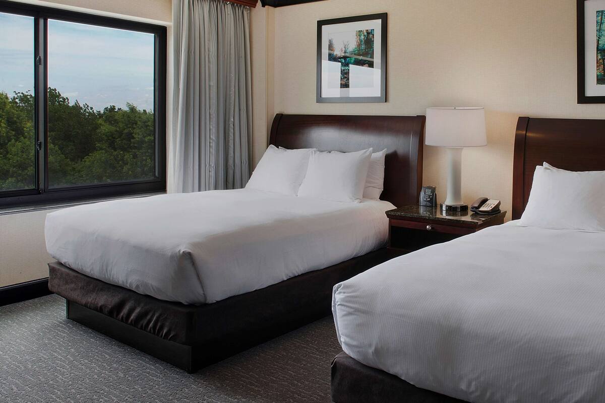 A hotel room with two beds, a large window with a view, a framed picture on the wall, and a bedside table with a lamp and phone.