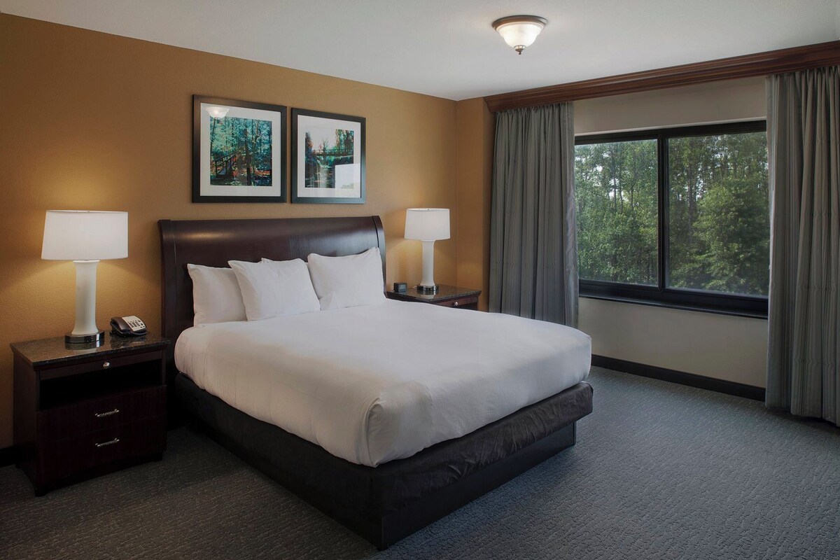 Habitación de hotel con una cama grande, dos mesitas de noche con lámparas, una mesita auxiliar, una ventana con cortinas y dos cuadros en la pared.