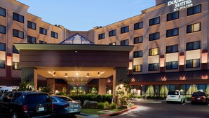 Un hotel DoubleTree Suites con diseño moderno, que cuenta con una entrada destacada con área cubierta y varios espacios de estacionamiento visibles.