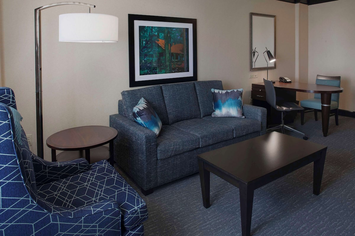 A hotel room with a gray sofa, a blue patterned armchair, a small round table, a coffee table, a desk with a chair, a lamp, and a framed painting on the wall.