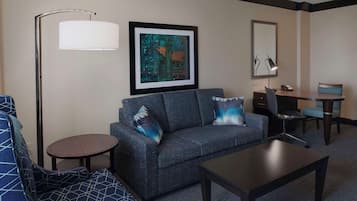 Quarto de hotel com sofá cinza, poltrona azul estampada, mesa pequena redonda, mesa de centro, escrivaninha com cadeira, luminária e quadro na parede.