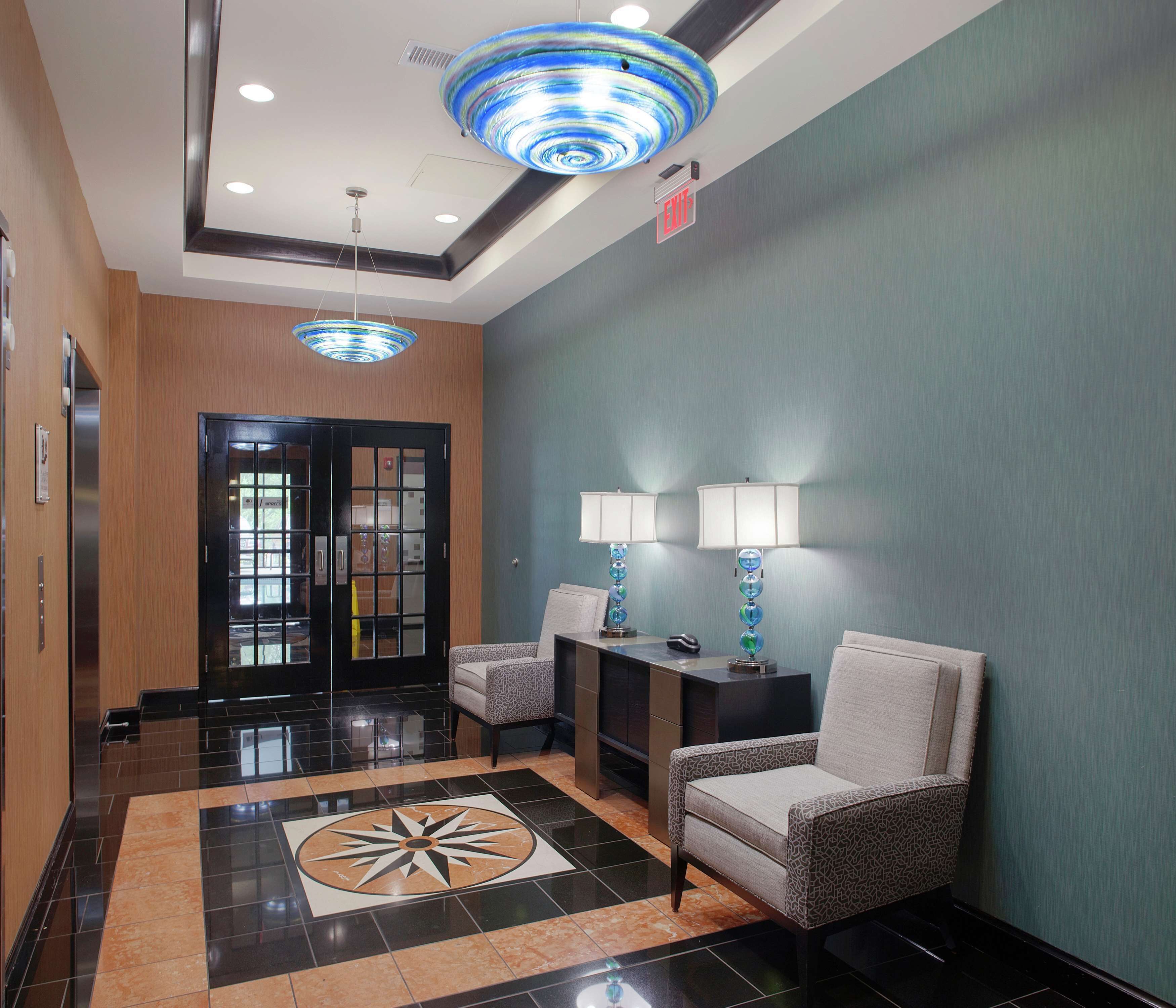 A lobby with a black and beige tiled floor, a blue and white patterned ceiling light, two chairs with lamps, and a door with a glass panel.