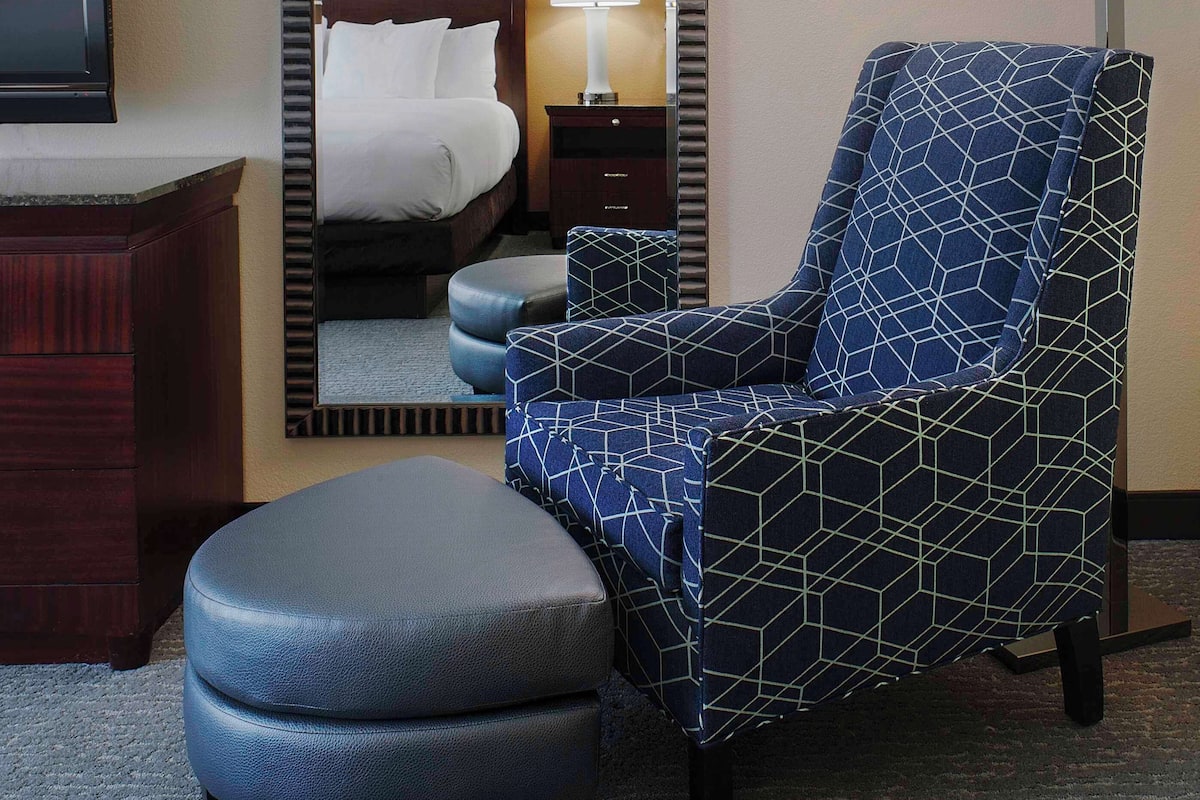 A hotel room with a bed, a chair, an ottoman, a desk, a lamp, and a television.