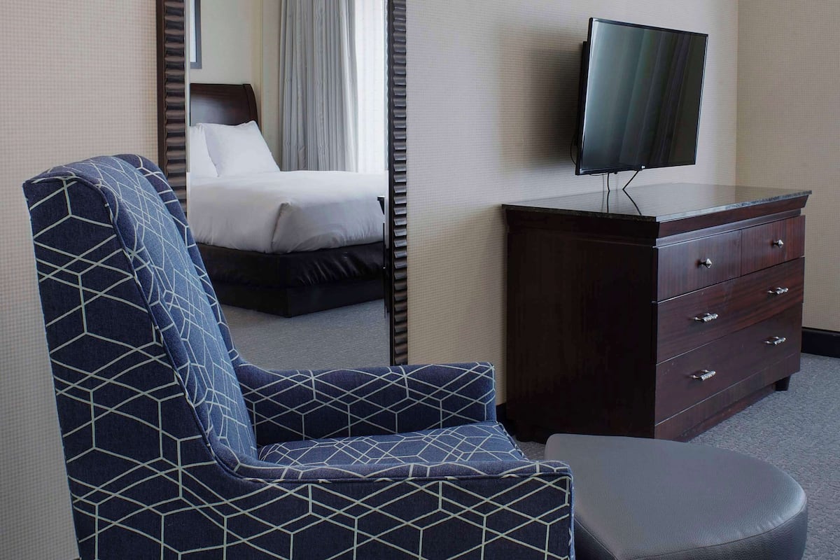 A hotel room with a bed, a TV on a dresser, a patterned armchair, and a small ottoman.