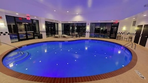 Una gran piscina interior con fondo iluminado en azul, rodeada por un suelo de baldosas y un borde de ladrillo.