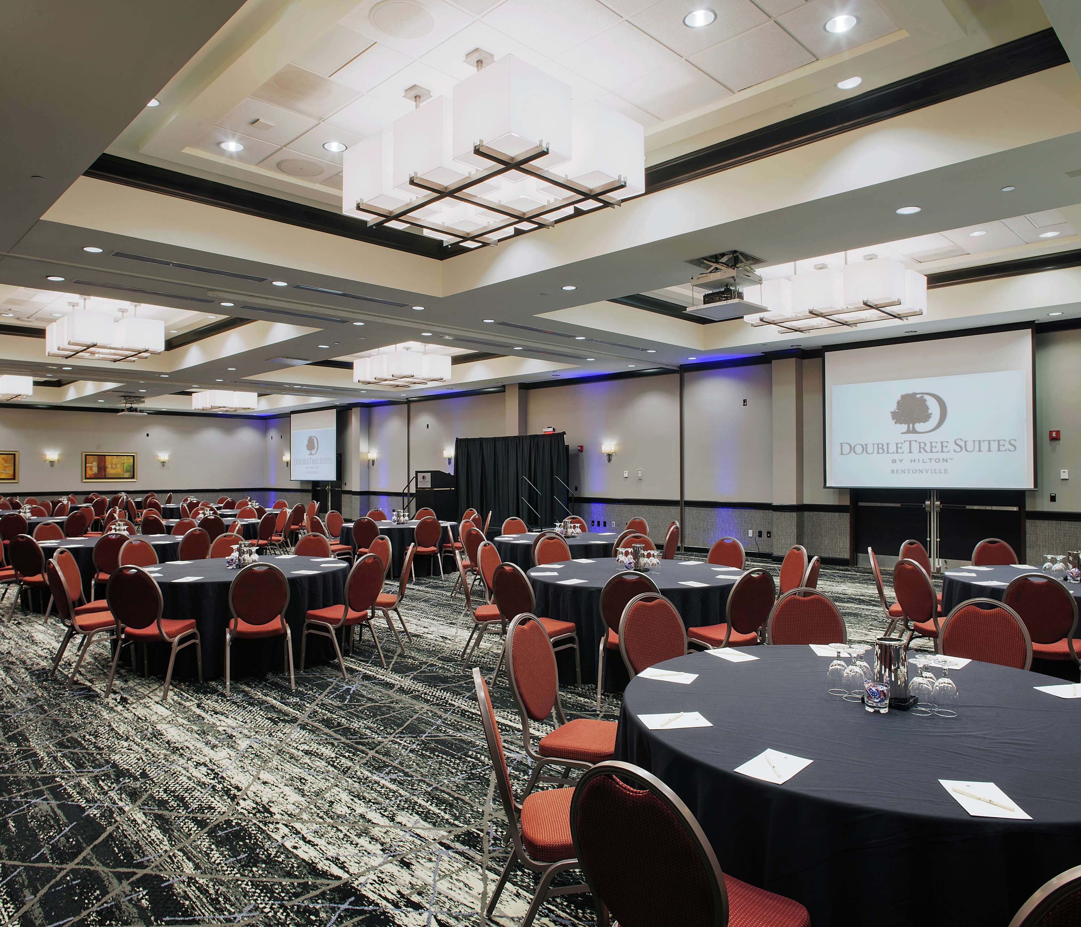 A conference room with multiple tables and chairs, a projector screen displaying "DOUBLETREE SUITES BY HILTON BENTONVILLE", and a stage with a podium.