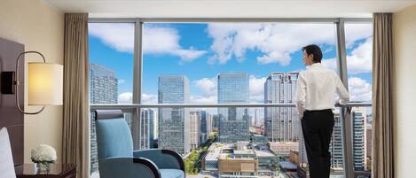 A hotel room with a large window, a bed, a chair, and a view of the cityscape.
