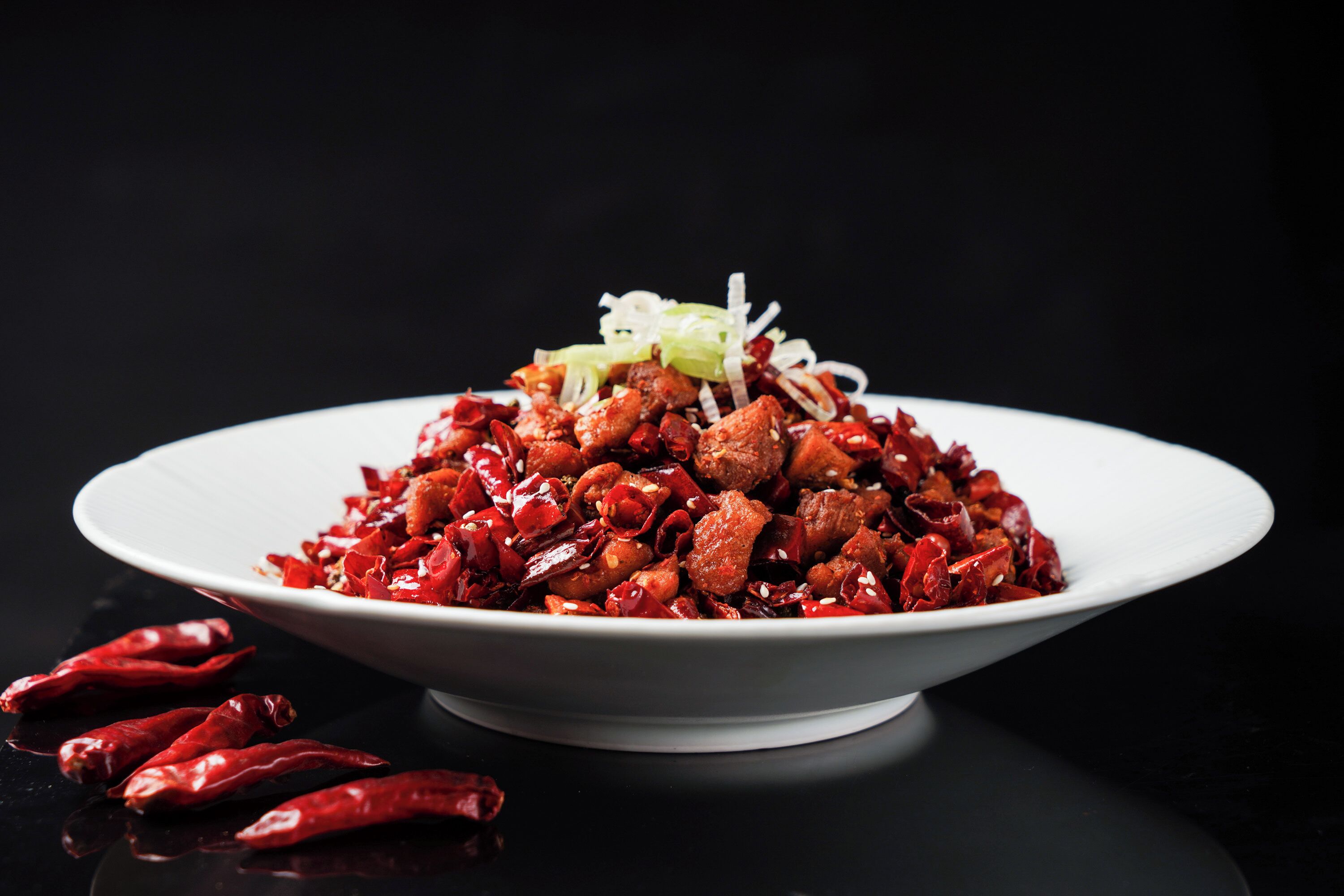 Un plato blanco con una pila de chiles rojos y carne, adornado con cebollas verdes, sobre una superficie negra.