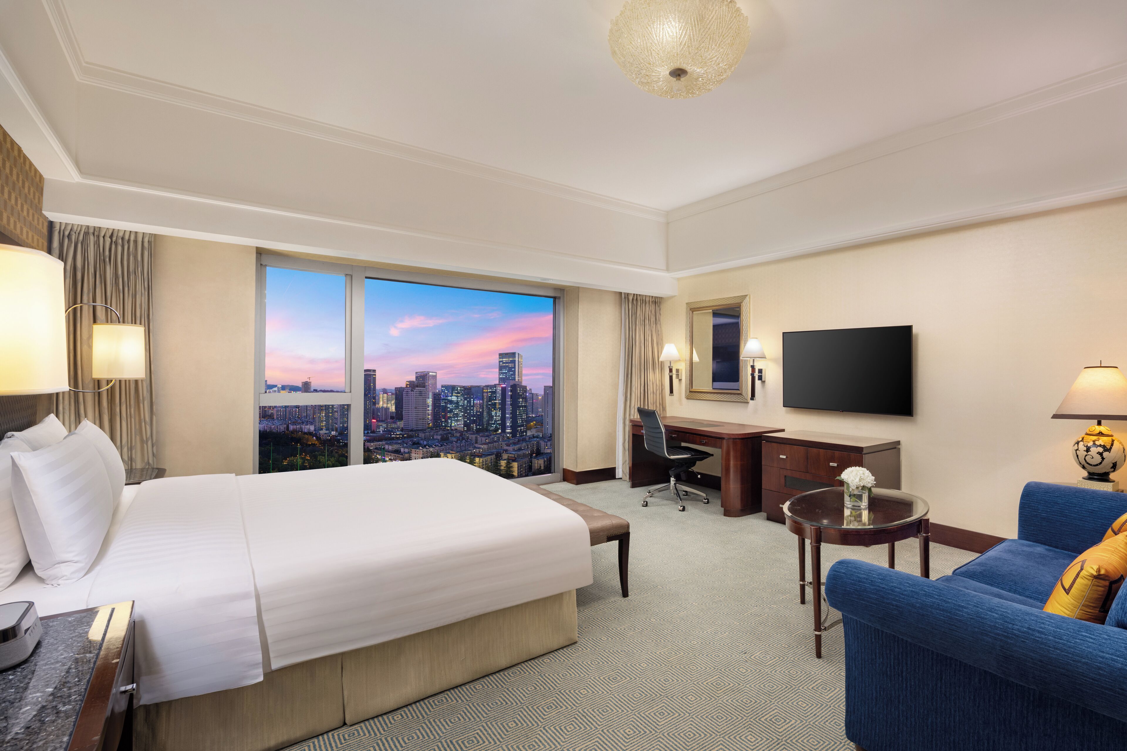 A hotel room with a large bed, a desk, a blue sofa, a TV, and a city view.