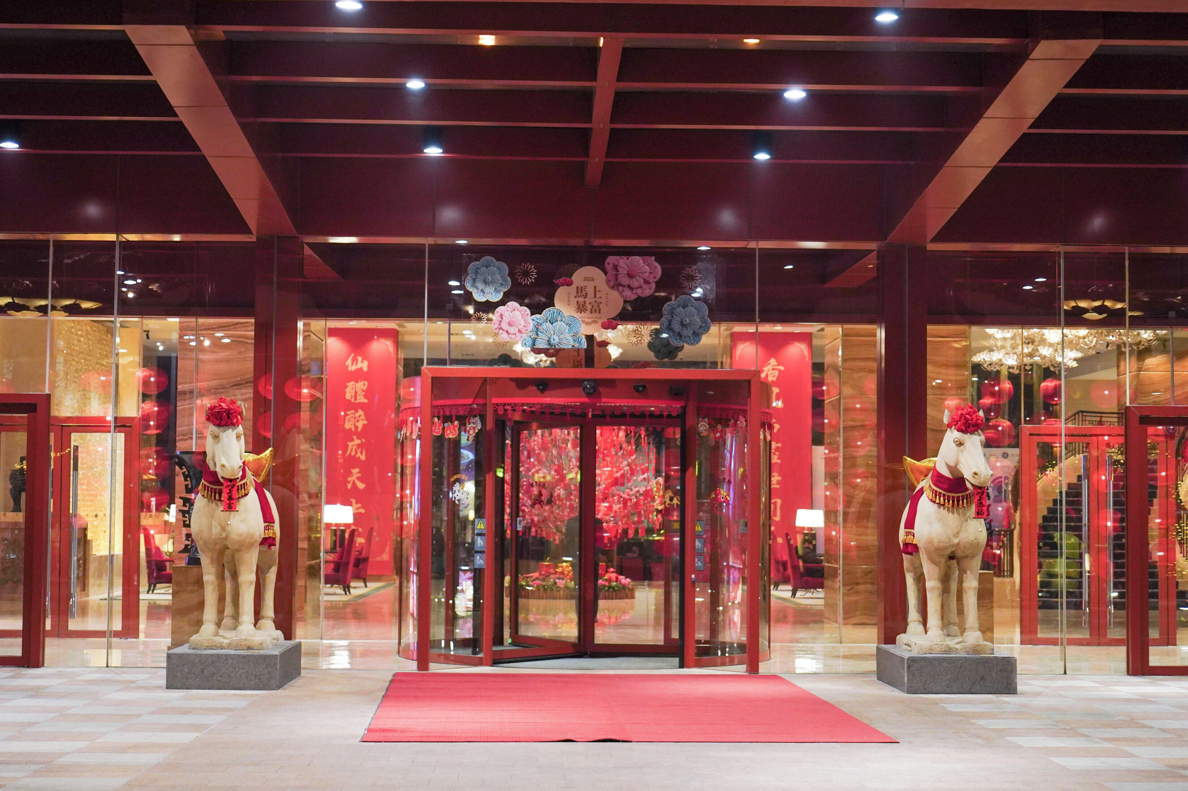 Entrada de un hotel con alfombra roja, puertas de cristal y dos estatuas ecuestres.