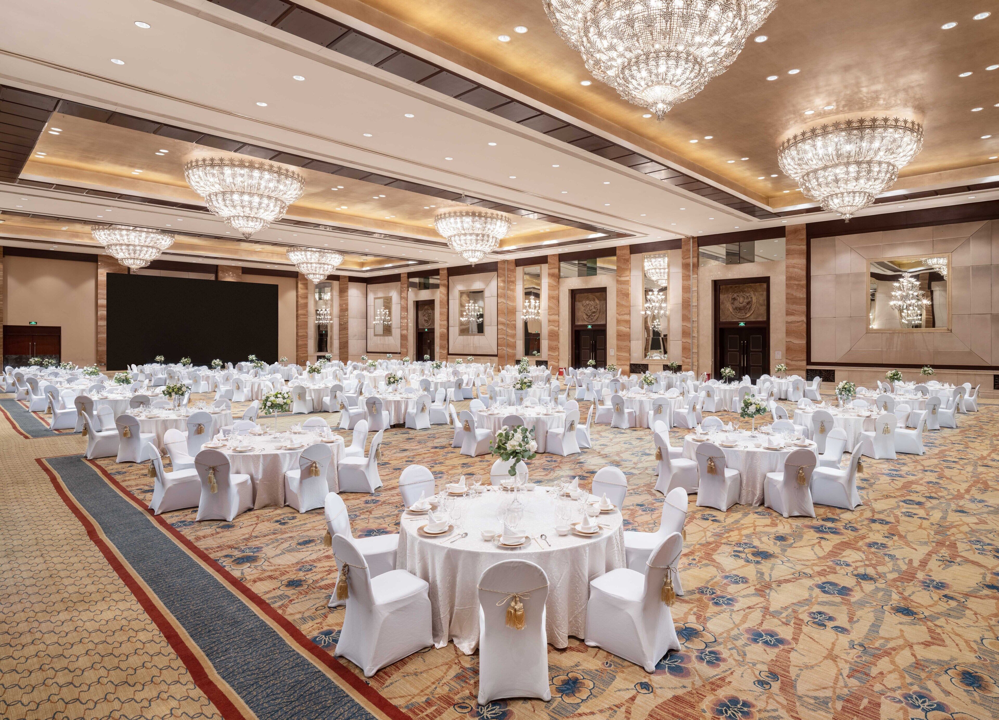 A large banquet hall with round tables and white chairs, chandeliers, and a carpeted floor.