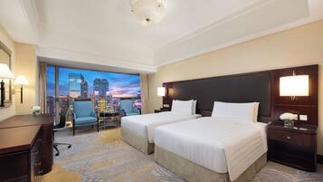 A hotel room with two beds, a desk, a chair, and a view of a cityscape at dusk.