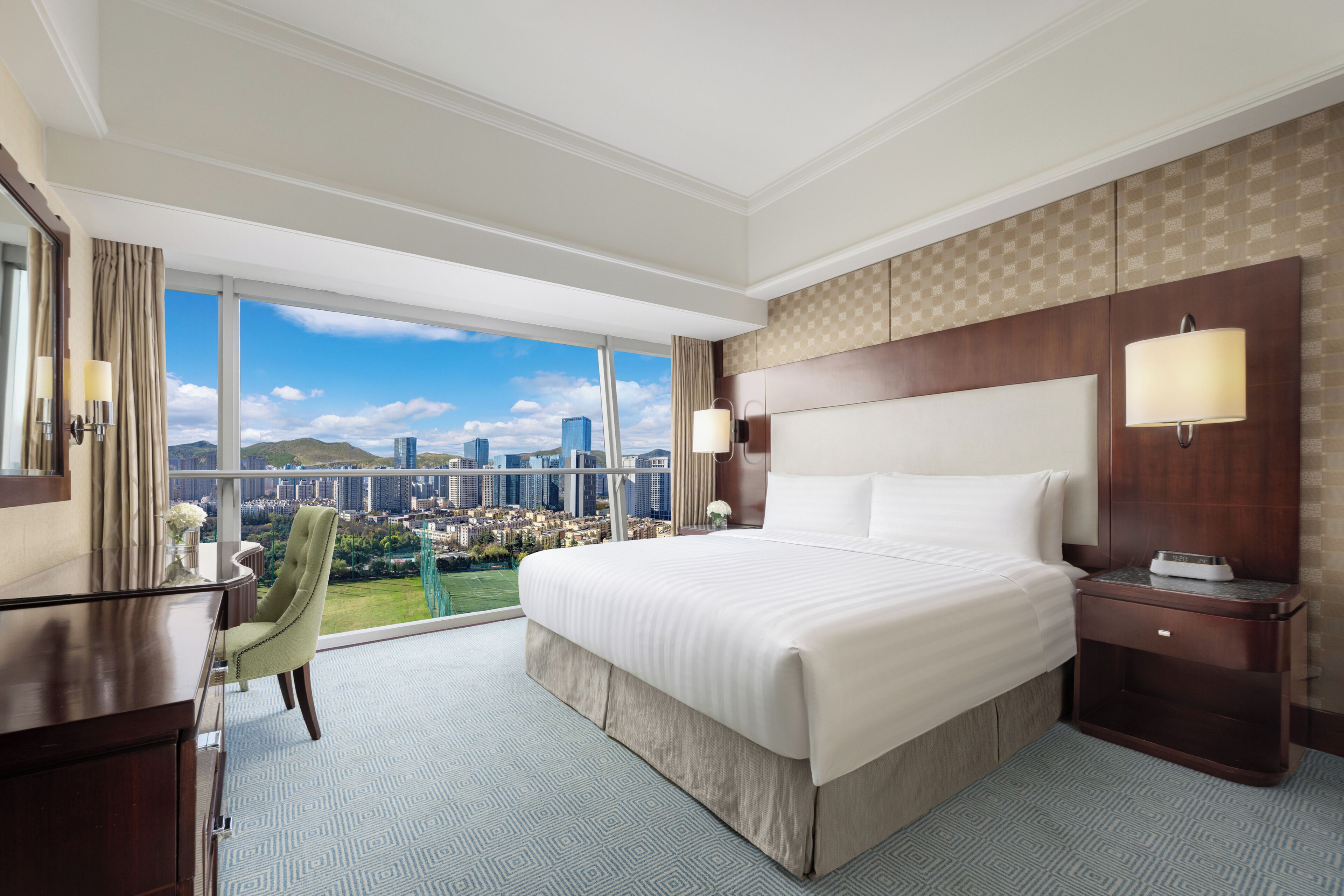 A hotel room with a large bed, a desk, a chair, a nightstand, and a view of a cityscape.