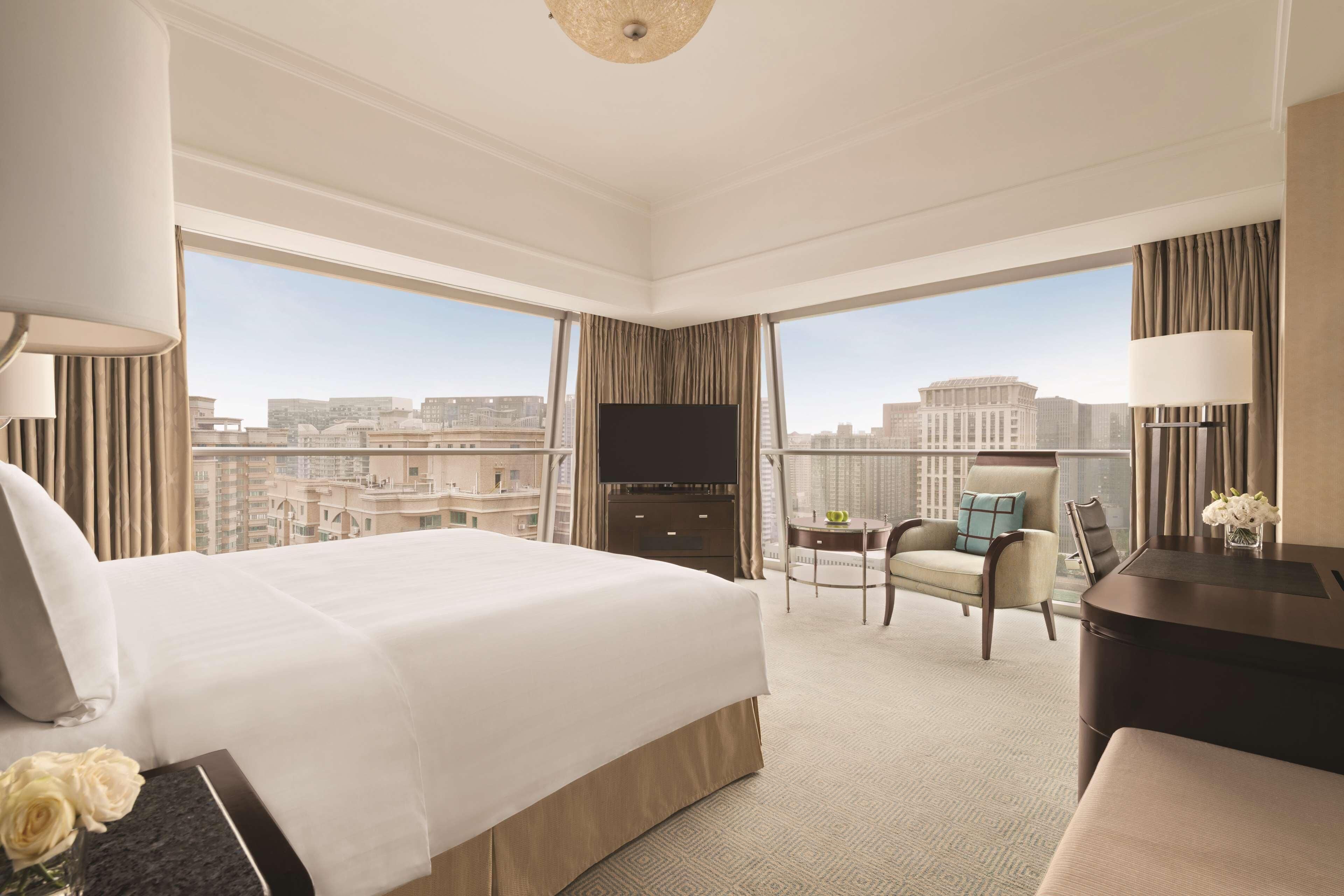 A hotel room with a large bed, a desk, a chair, a TV, and a view of the cityscape.