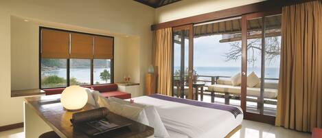 Quarto de hotel com uma cama grande, uma escrivaninha com abajur, vista para o mar e uma varanda com área de estar ao ar livre.