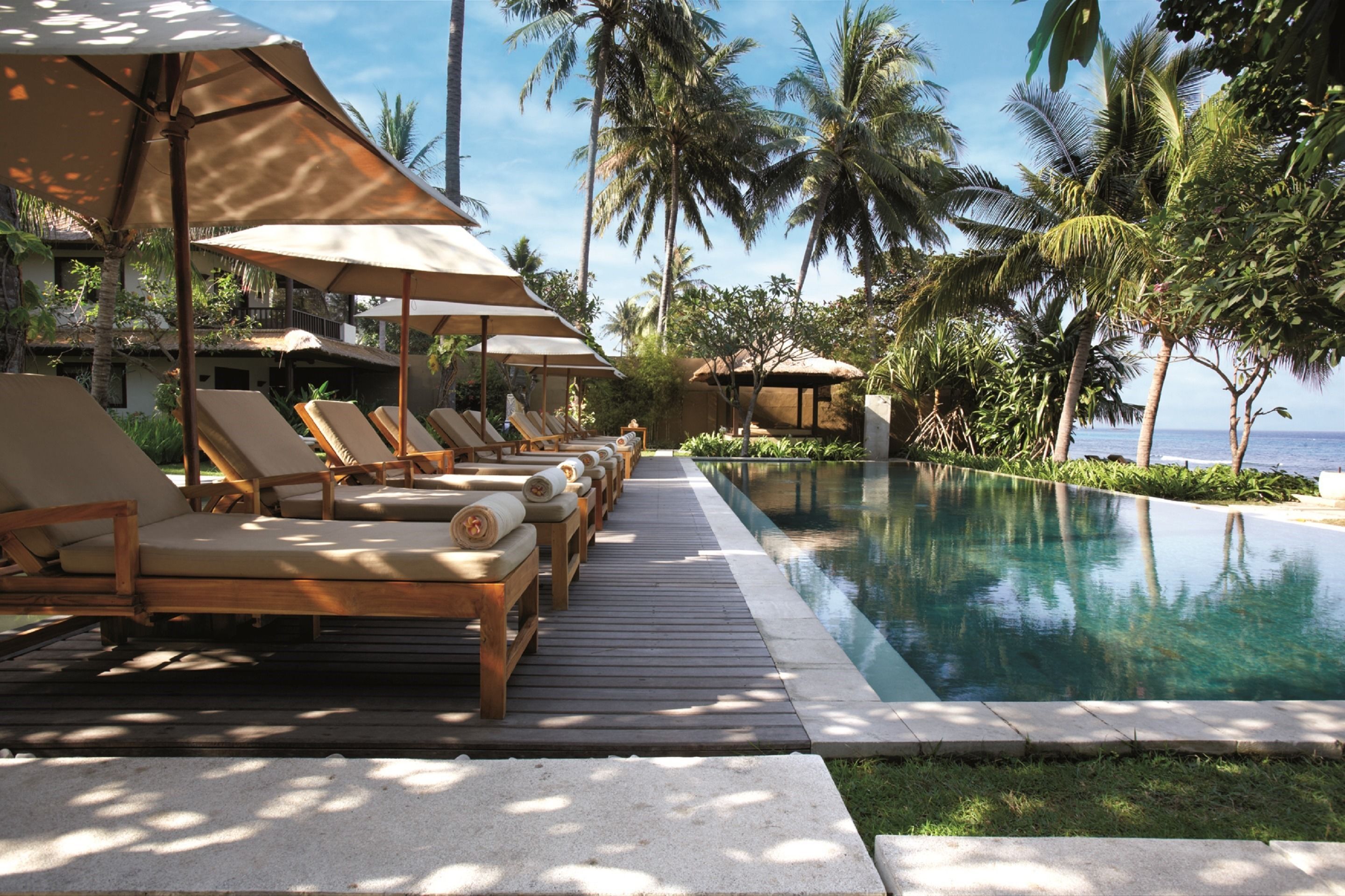 A poolside area with lounge chairs and umbrellas, surrounded by palm trees and a clear view of the ocean.