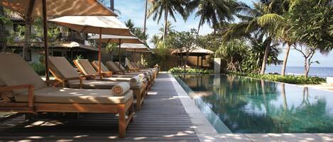 A poolside area with lounge chairs and umbrellas, surrounded by palm trees and a clear view of the ocean.