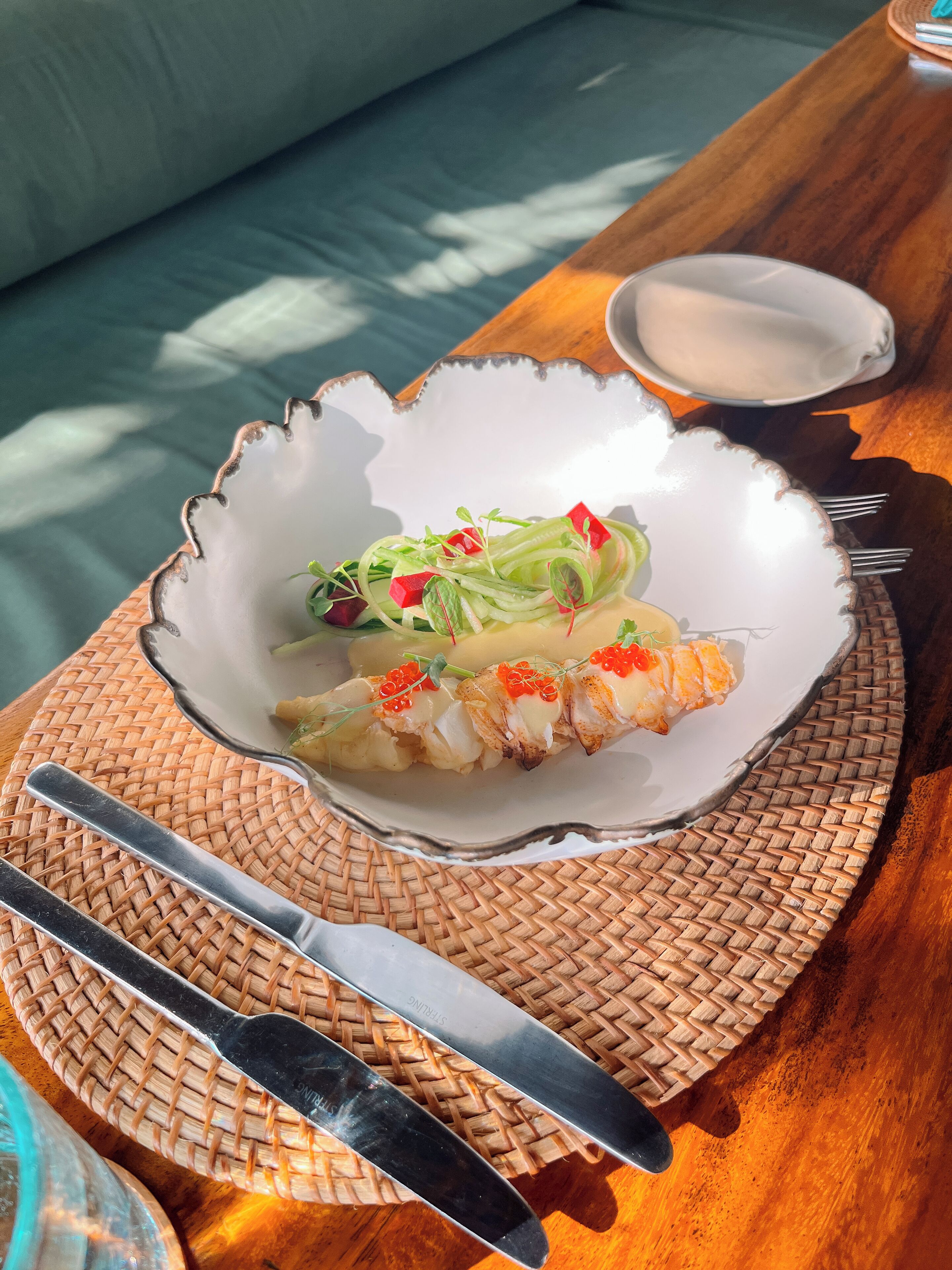 A plate of sushi with a decorative bowl on a woven mat, accompanied by a knife and fork on a wooden table.
