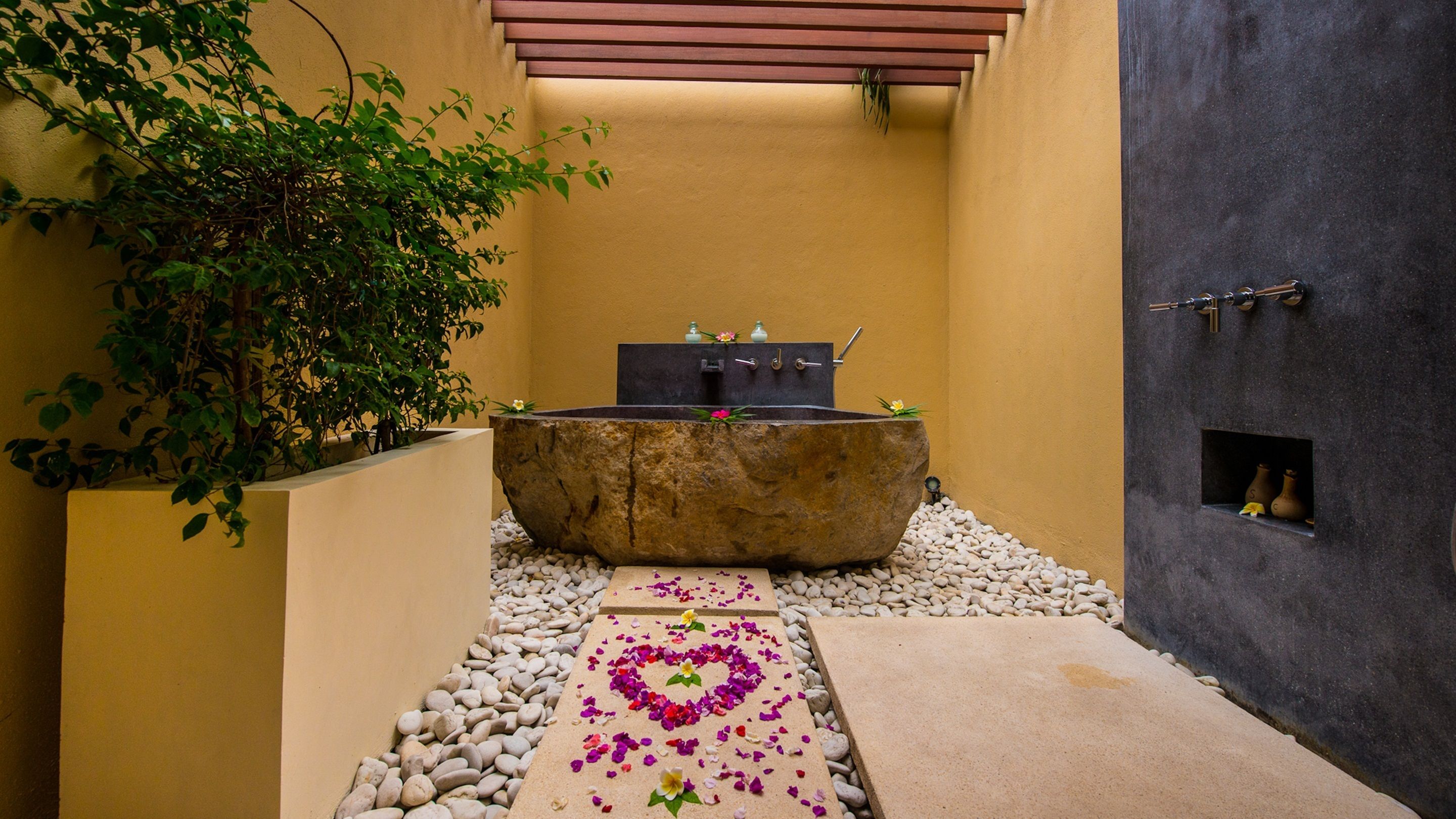 A stone bathtub in a narrow, yellow-walled corridor with a pebble path leading to it, surrounded by greenery and a small window with vases.
