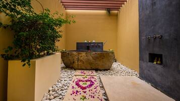 A stone bathtub in a narrow, yellow-walled corridor with a pebble path leading to it, surrounded by greenery and a small window with vases.