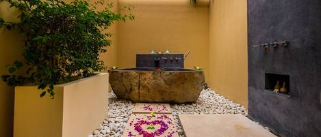 A stone bathtub in a narrow, yellow-walled corridor with a pebble path leading to it, surrounded by greenery and a small window with vases.