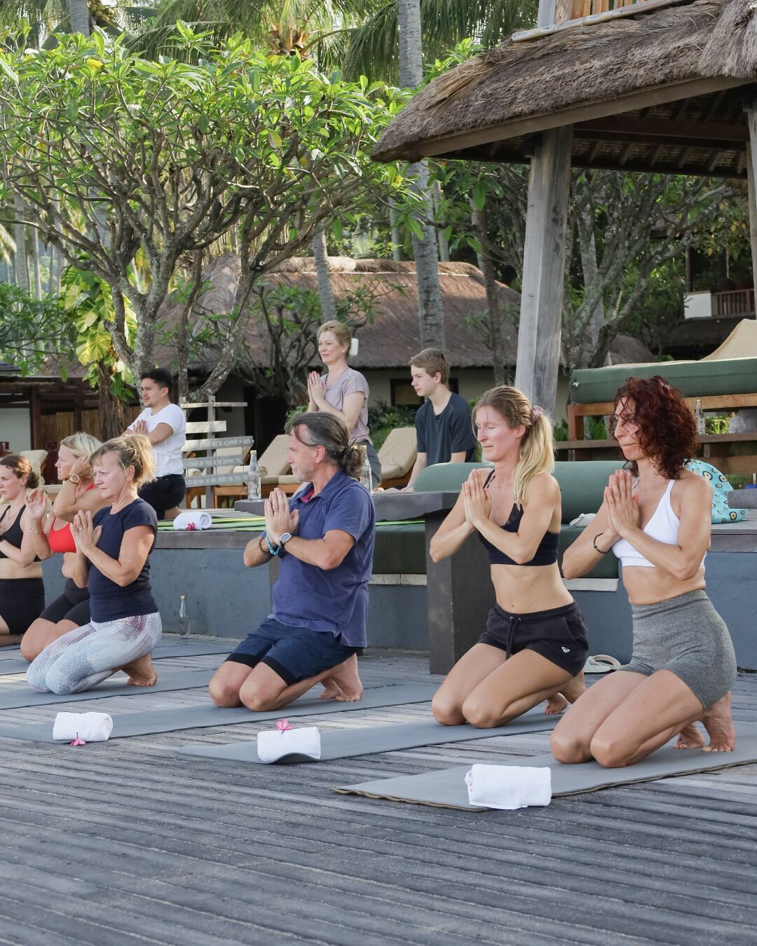 A group of people performing yoga on a wooden deck with a thatched roof structure in the background.