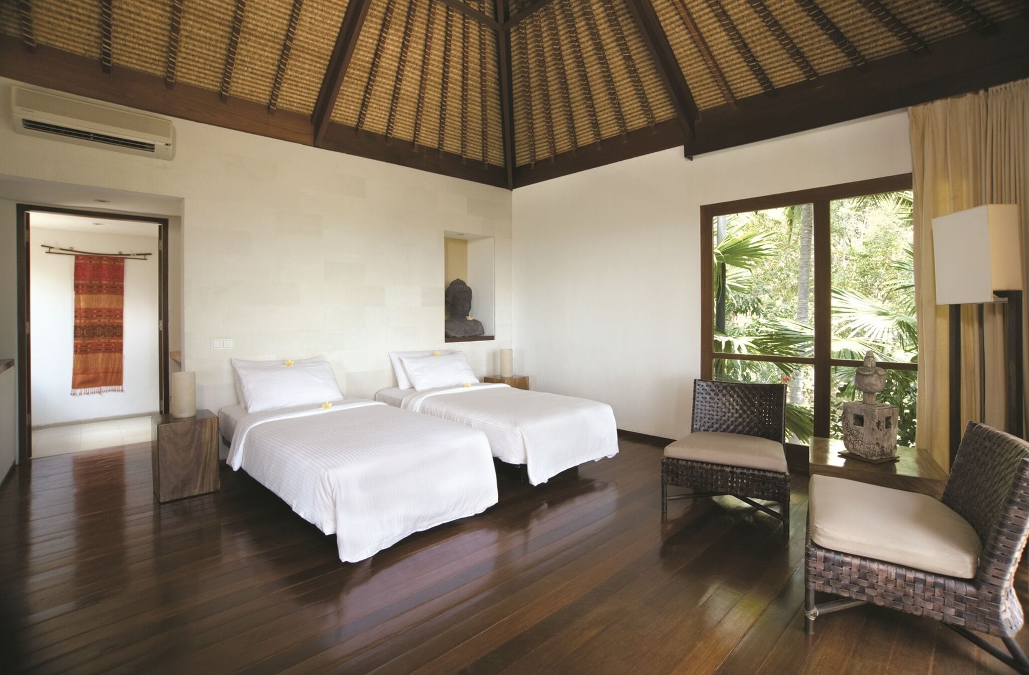 A hotel room with two beds, a sitting area with a chair, a large window with a view of greenery, and a ceiling with a woven pattern.