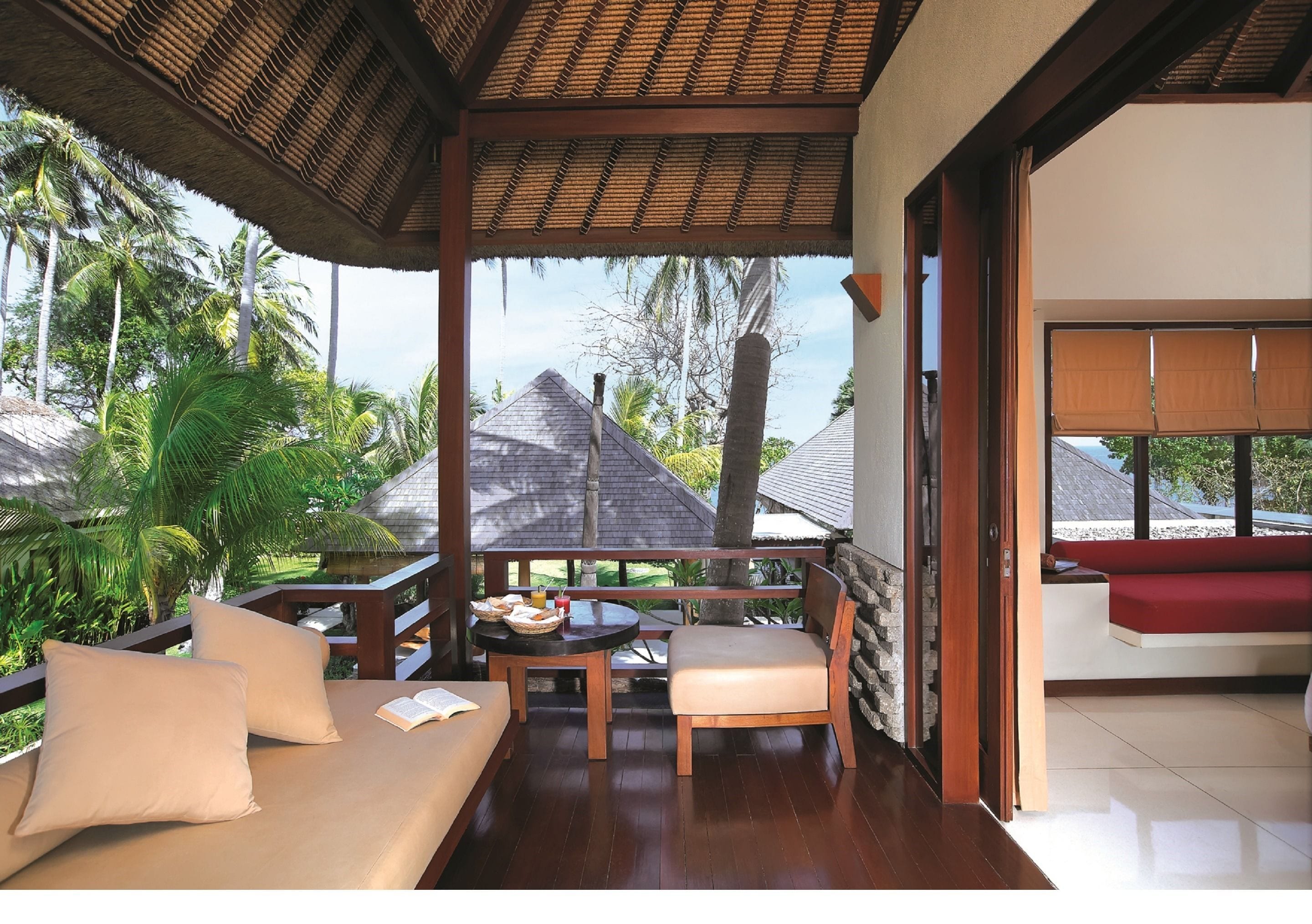 A covered outdoor seating area with a wooden bench and cushions, a table with a book and a basket, and a view of palm trees and a house in the background.