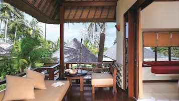 A covered outdoor seating area with a wooden bench and cushions, a table with a book and a basket, and a view of palm trees and a house in the background.