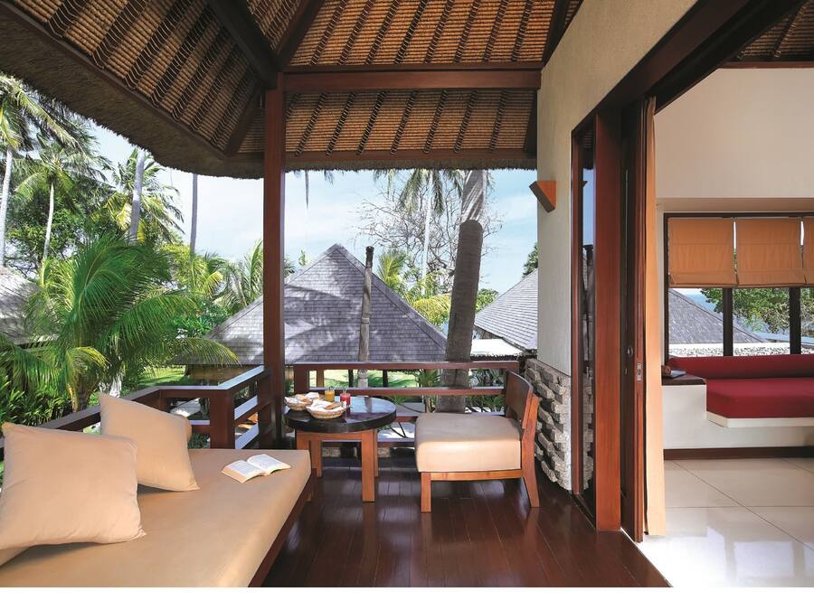 A covered outdoor seating area with a wooden bench and cushions, a table with a book and a basket, and a view of palm trees and a house in the background.