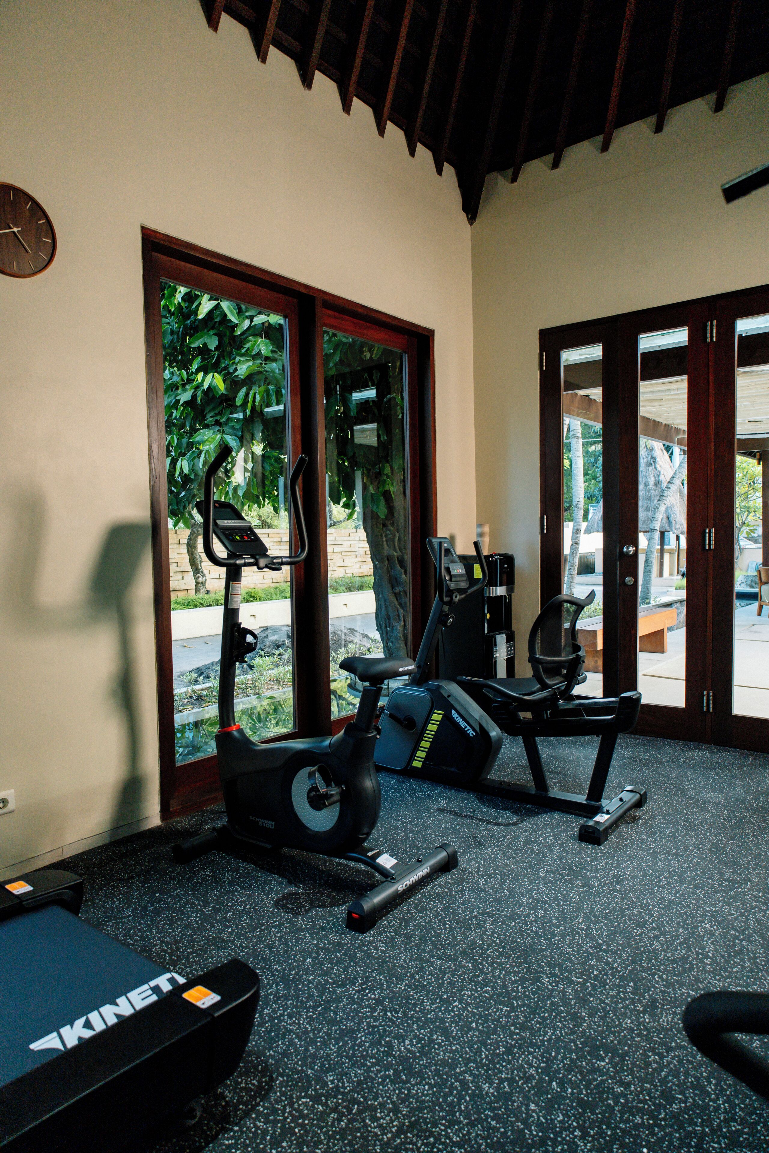 A gym with stationary bikes and a treadmill, large windows with a view of greenery, and a wooden ceiling.