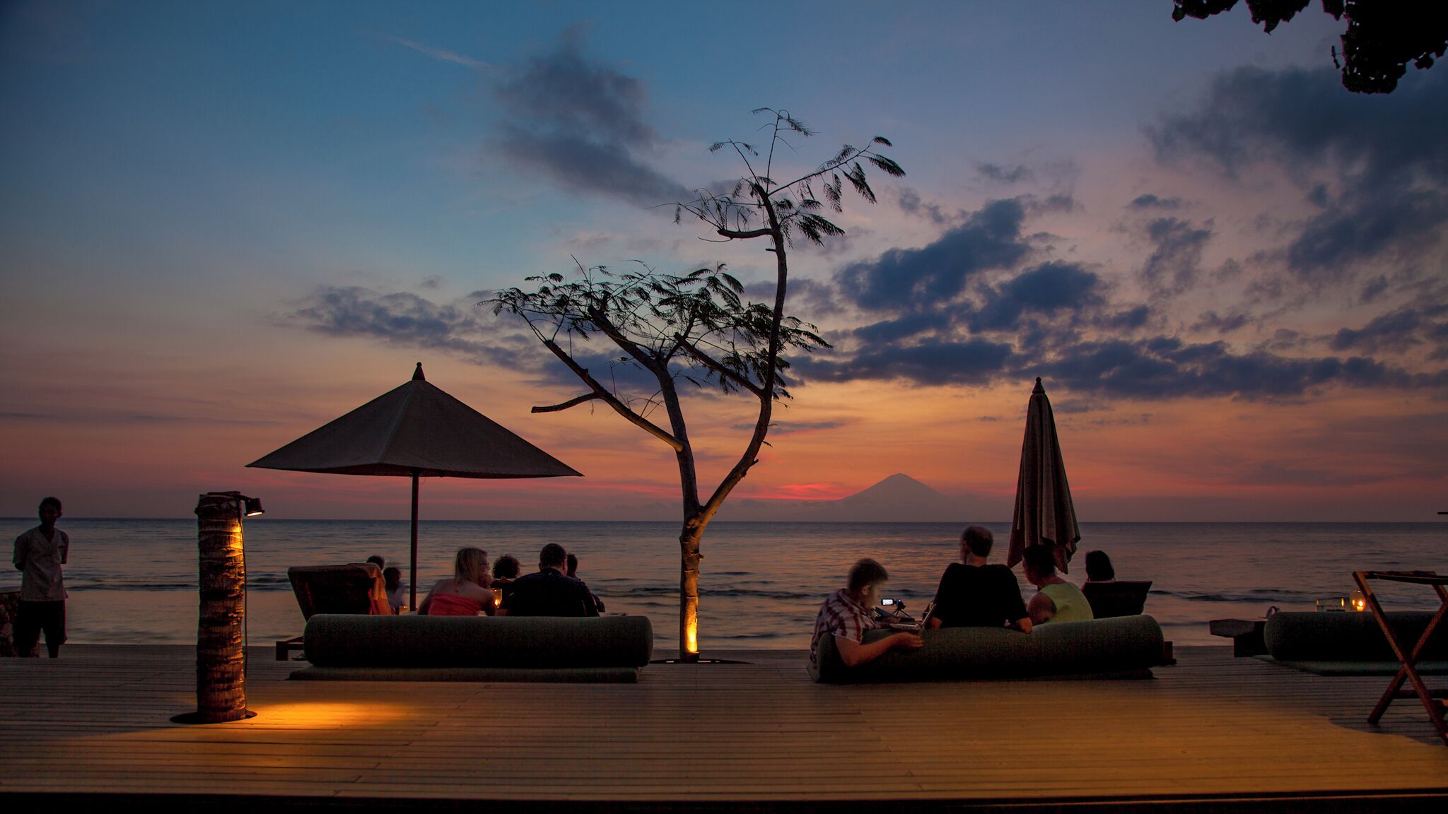 A sunset view from a wooden deck with lounge chairs and umbrellas, overlooking the ocean.