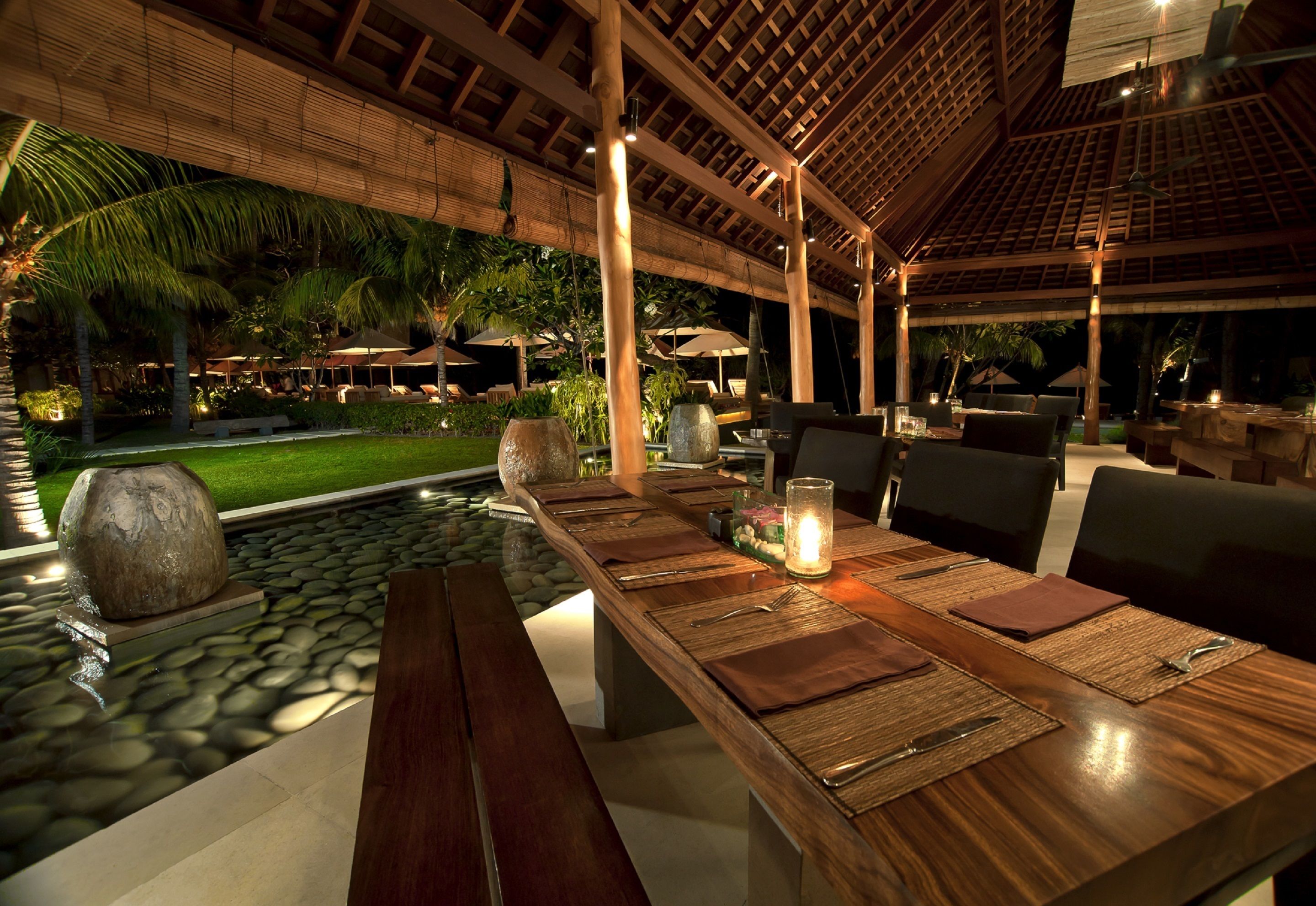A dining area with a long wooden table set for a meal, surrounded by chairs, with a stone-lined pool and palm trees in the background.